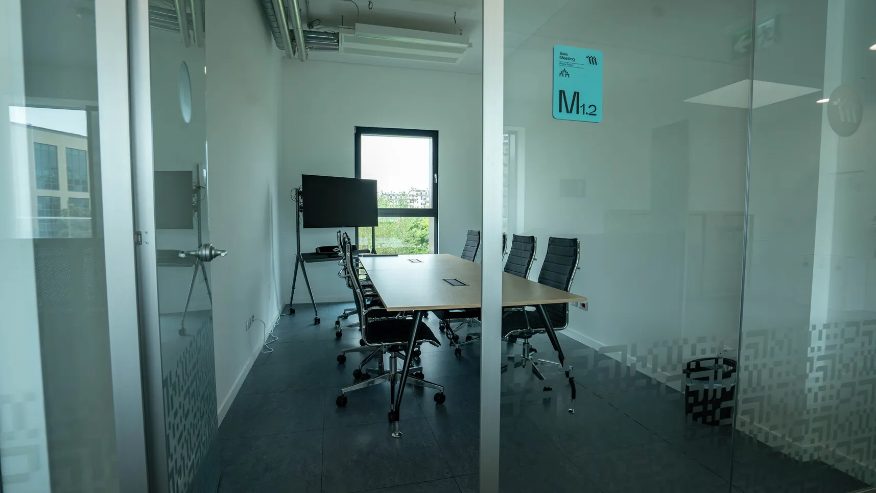 Sala riunioni moderna con tavolo rettangolare, sei sedie girevoli nere e un grande schermo davanti alla finestra.