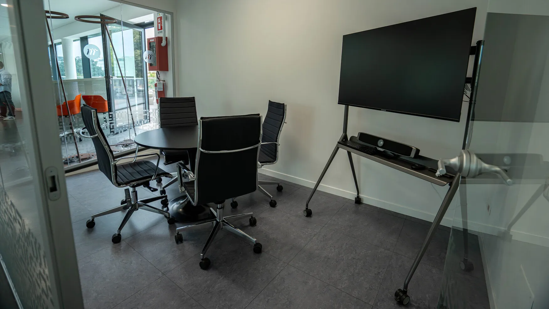 Sala riunioni moderna con tavolo rotondo, quattro sedie nere girevoli e grande schermo su cavalletto.