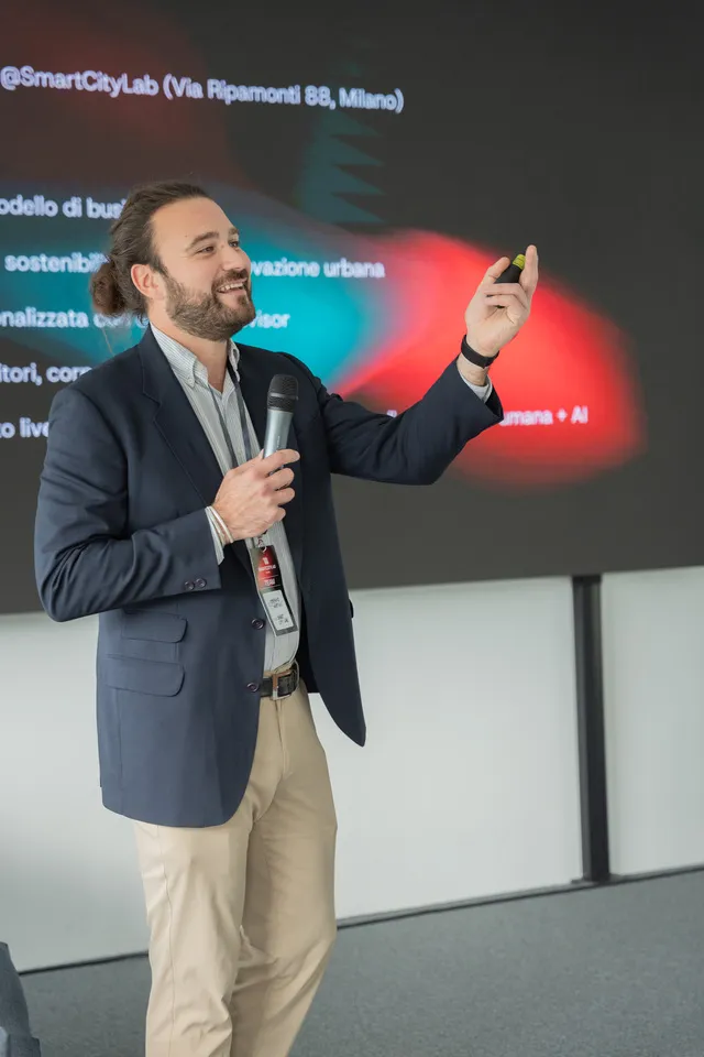 Uomo con giacca blu e pantaloni beige tiene un microfono e presenta davanti a uno schermo con testo e grafica colorata.