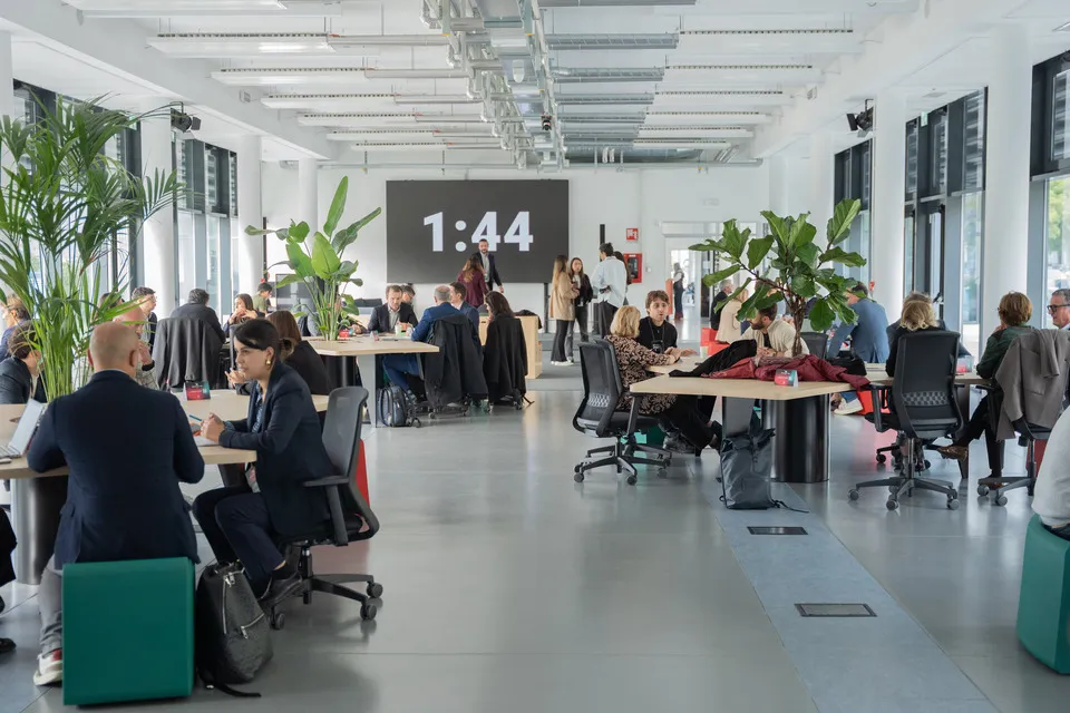 Spazio di coworking moderno con persone che lavorano e conversano attorno a grandi tavoli con piante, illuminato da ampie finestre.