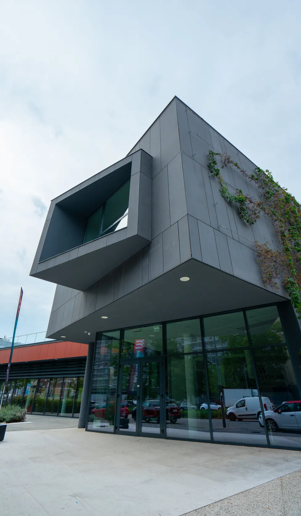 Edificio moderno con facciata grigia e grandi finestre riflettenti all'ingresso, piante rampicanti sulla parete.