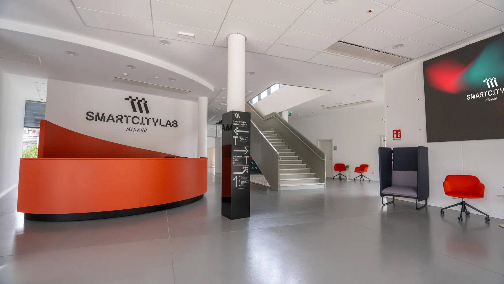 Reception con bancone arancione in un ambiente moderno con logo Smart City Lab Milano, poltrone rosse e scala interna.