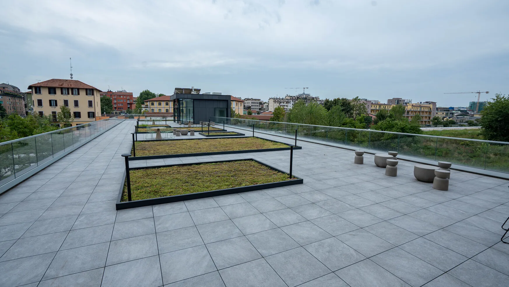 Ampio terrazzo piastrellato con aiuole verdi rialzate, sedute moderne in pietra, ringhiere di vetro e vista su edifici residenziali.