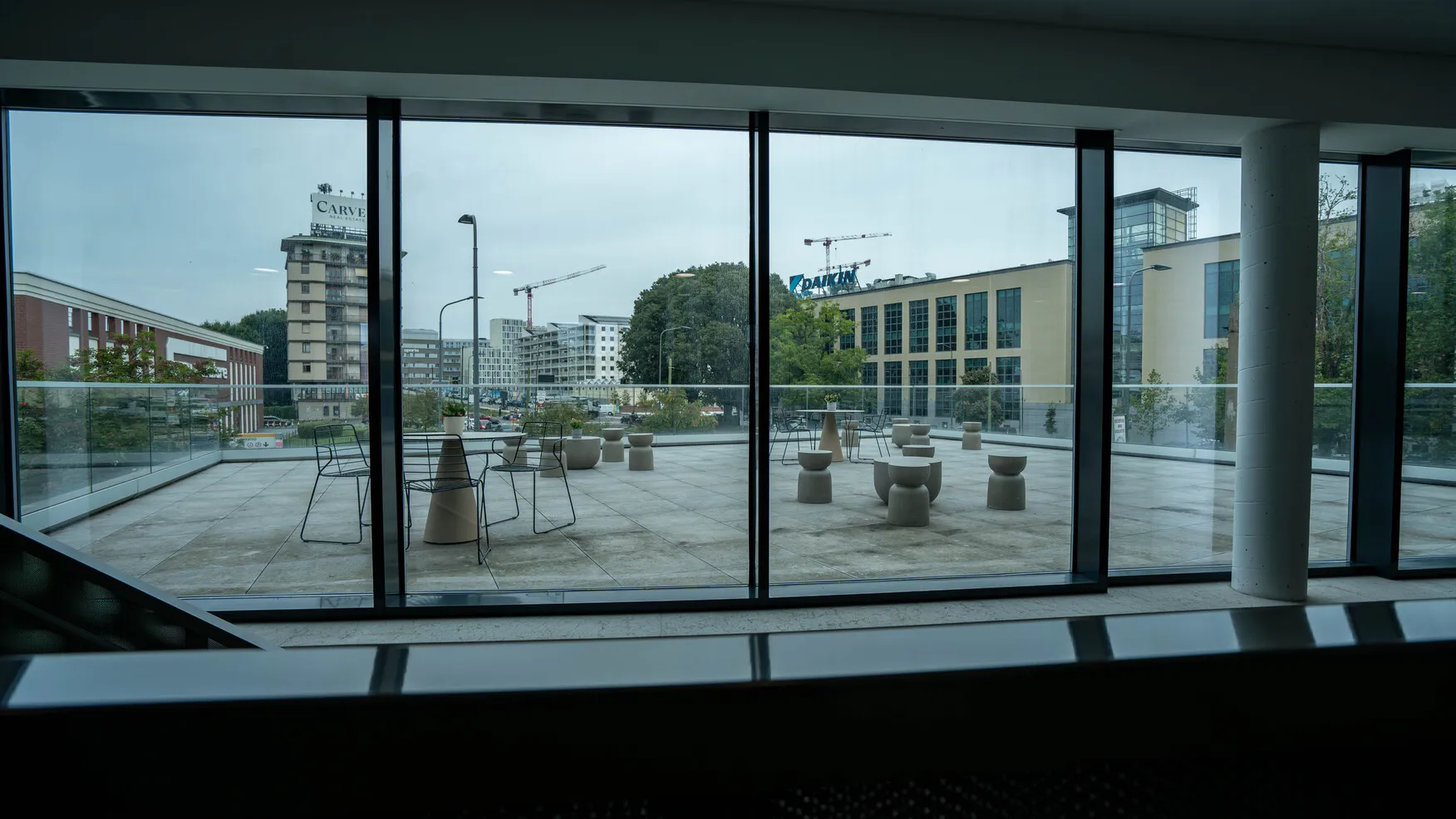 Terrazza con sedie e tavolini all'aperto vista attraverso grandi finestre, sullo sfondo edifici urbani e gru da costruzione.