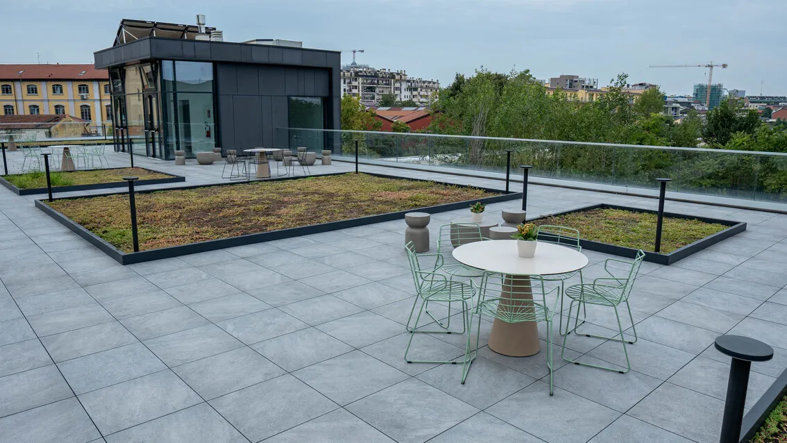 Terrazza panoramica con pavimento grigio, tavolo rotondo bianco con quattro sedie in metallo verde, aiuole piantumate e parapetto in vetro.