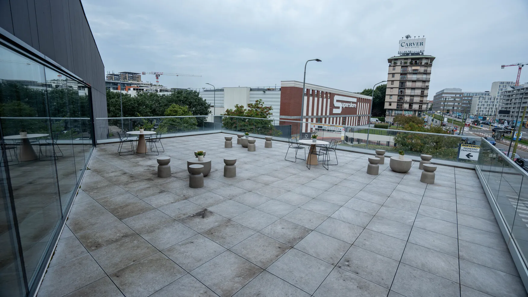 Terrazza privata sul tetto con sedie, tavoli tondi e sgabelli grigi, vista su edificio commerciale con insegna Superstore e strada urbana.