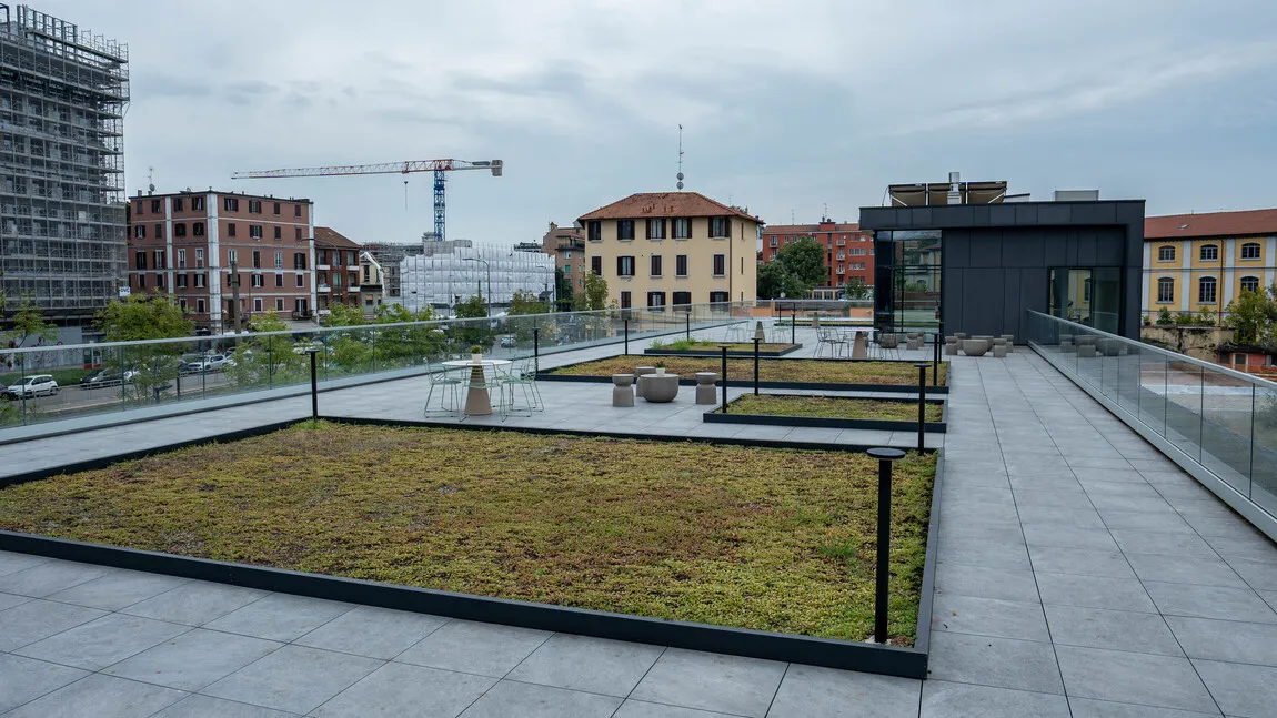 Terrazza sul tetto con aiuole erbose, tavoli e sedie in metallo, circondata da edifici urbani e gru da costruzione sullo sfondo.
