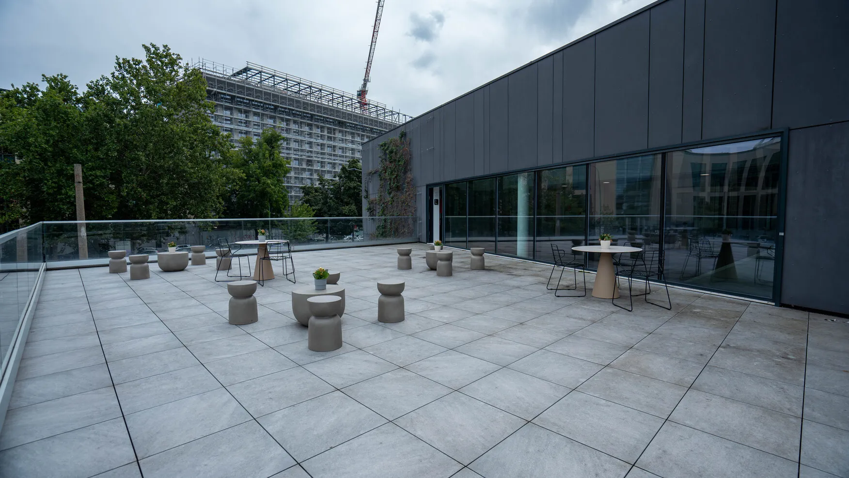 Ampia terrazza con pavimento in piastrelle grigie, arredi moderni beige e neri, decorazioni floreali e vista su alberi e un edificio in costruzione.