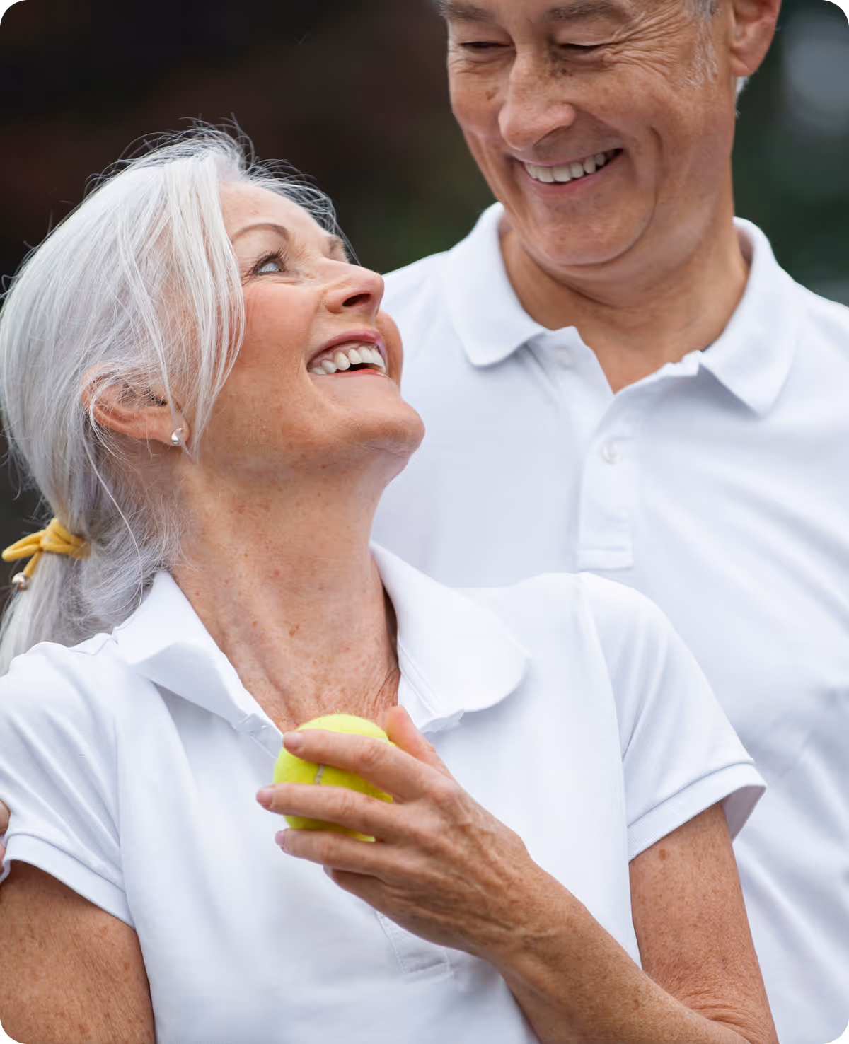 Smiling elderly woman holding a tennis ball looking up at a smiling elderly man in white polo shirts.