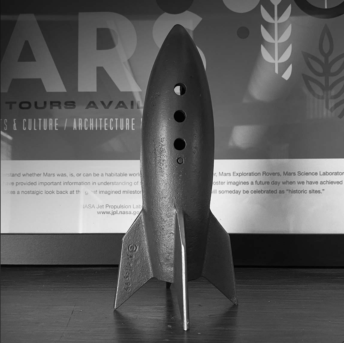 Black metal rocket model with three fins and three circular windows standing on a dark surface in front of a Mars exploration poster.
