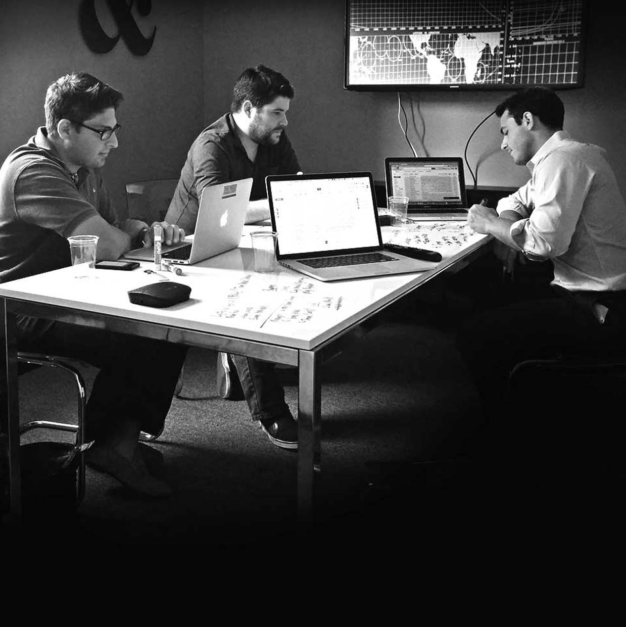 Three men working on laptops around a white table with notes, with a world map displayed on a screen in the background.