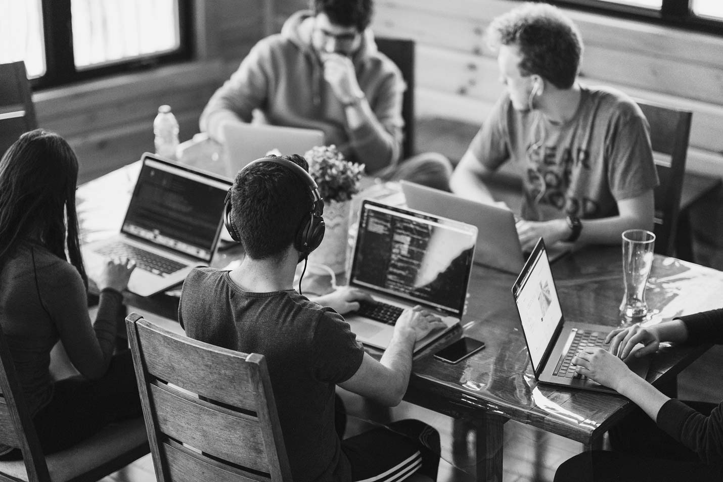 Group of five people working on laptops around a wooden table in a casual setting.