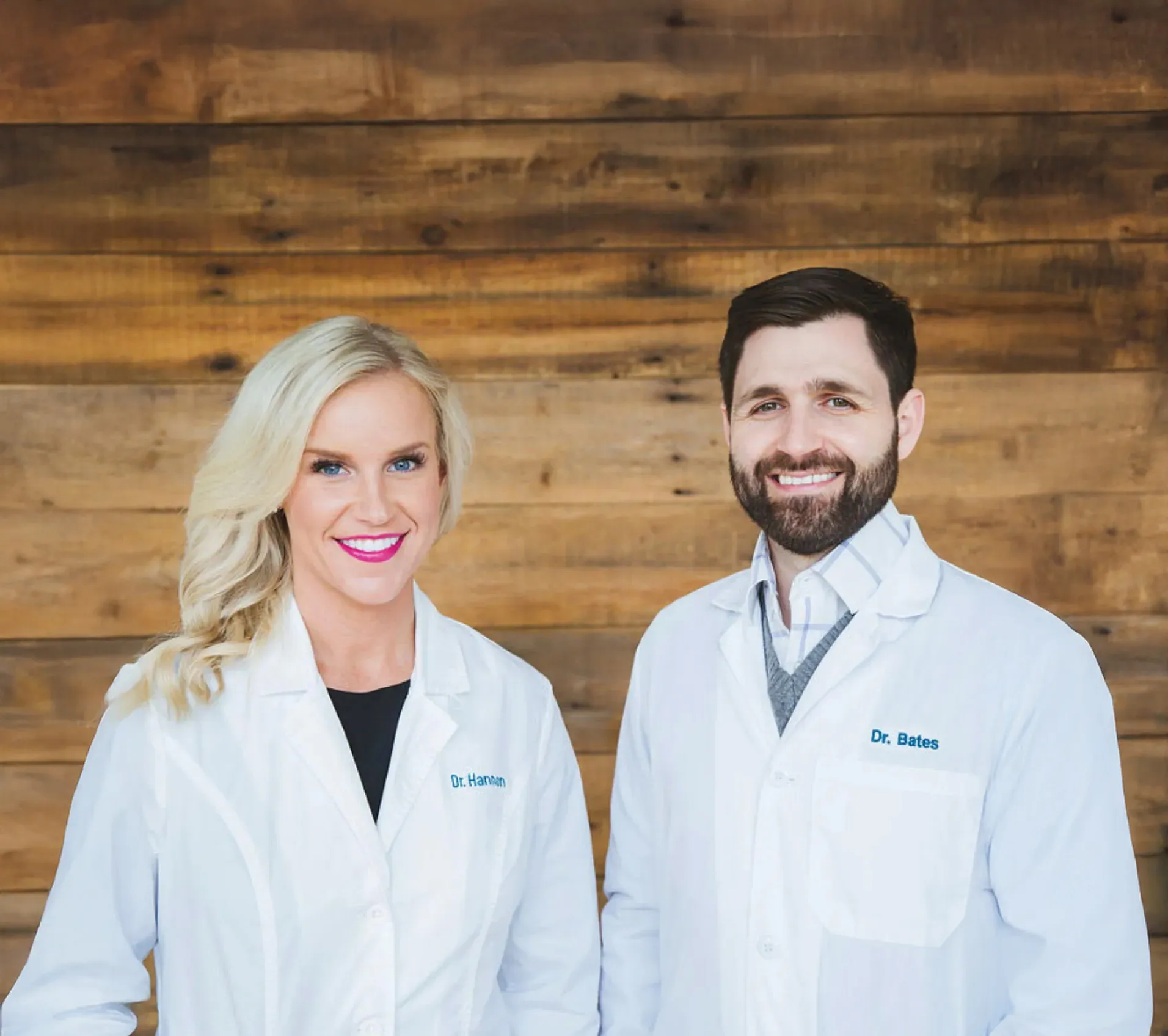 Dr. David Bates and Dr. Katie Hannon smiling in white lab coats, standing against a rustic wooden wall backdrop.