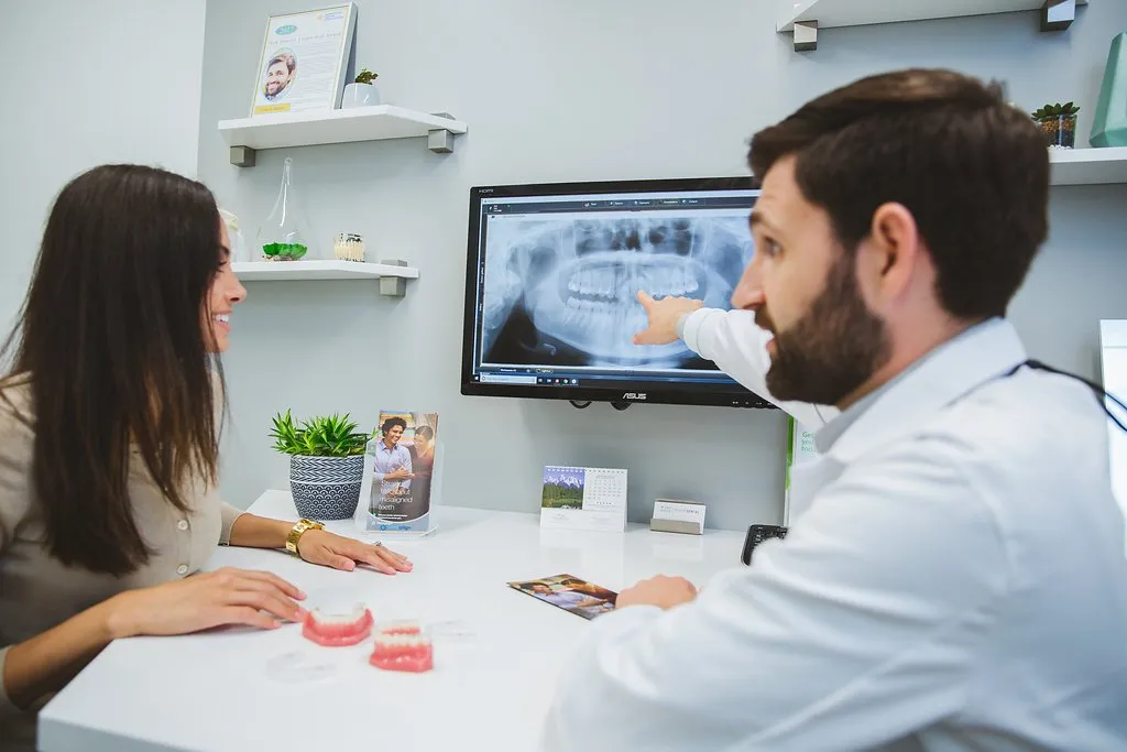 A dentist reviewing a panoramic dental X-ray on a monitor with a patient during a consultation, using models to explain treatment options.