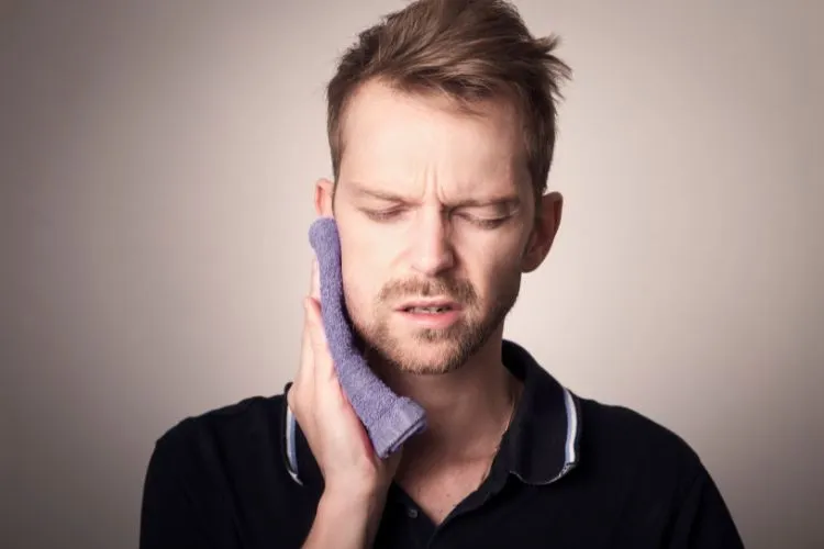A young man pressing an ice pack to his cheek with eyes closed, showing visible discomfort from dental pain or swelling.
