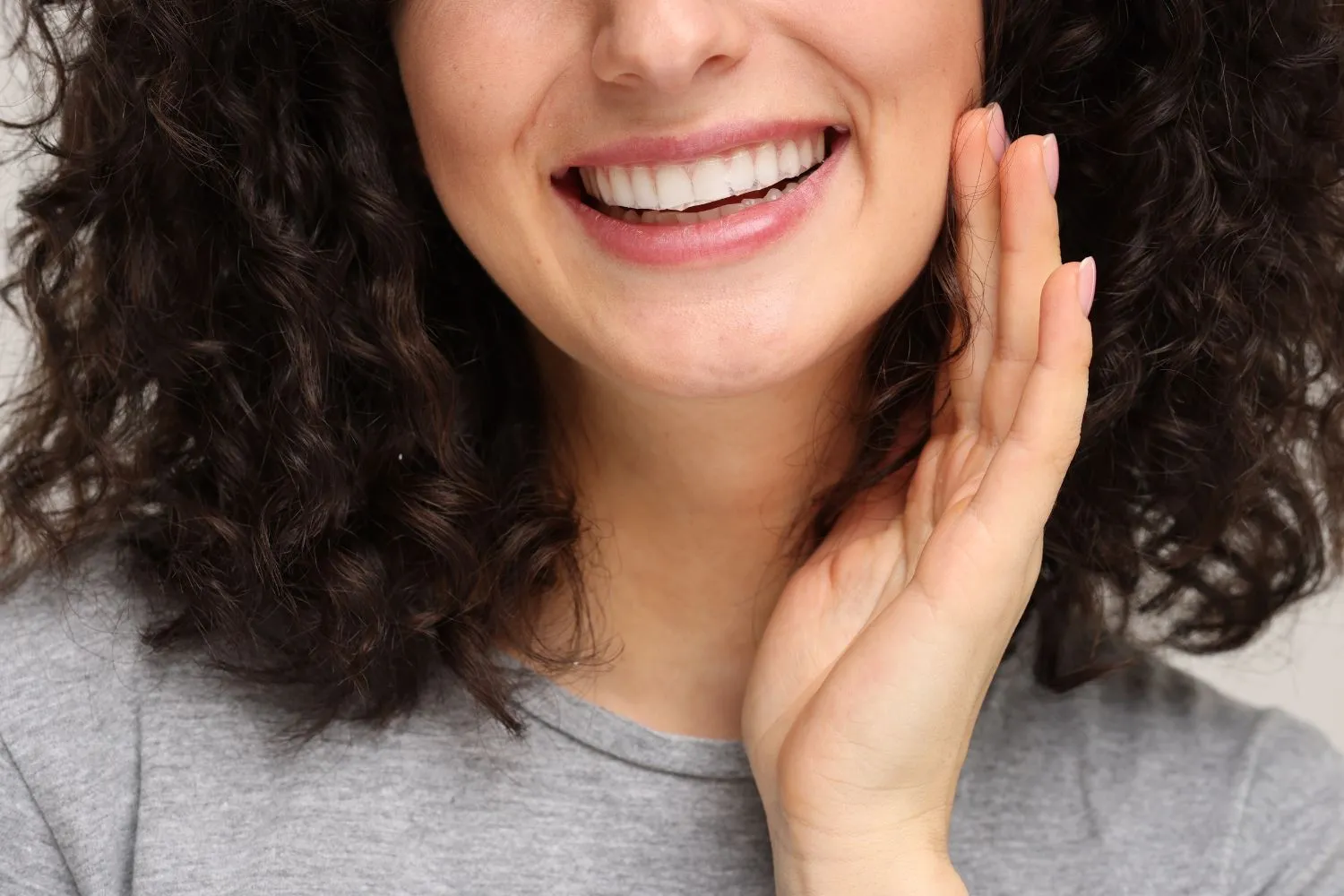 Close-up of a woman with curly hair smiling brightly and touching her cheek, showcasing straight, white teeth after cosmetic dental care.