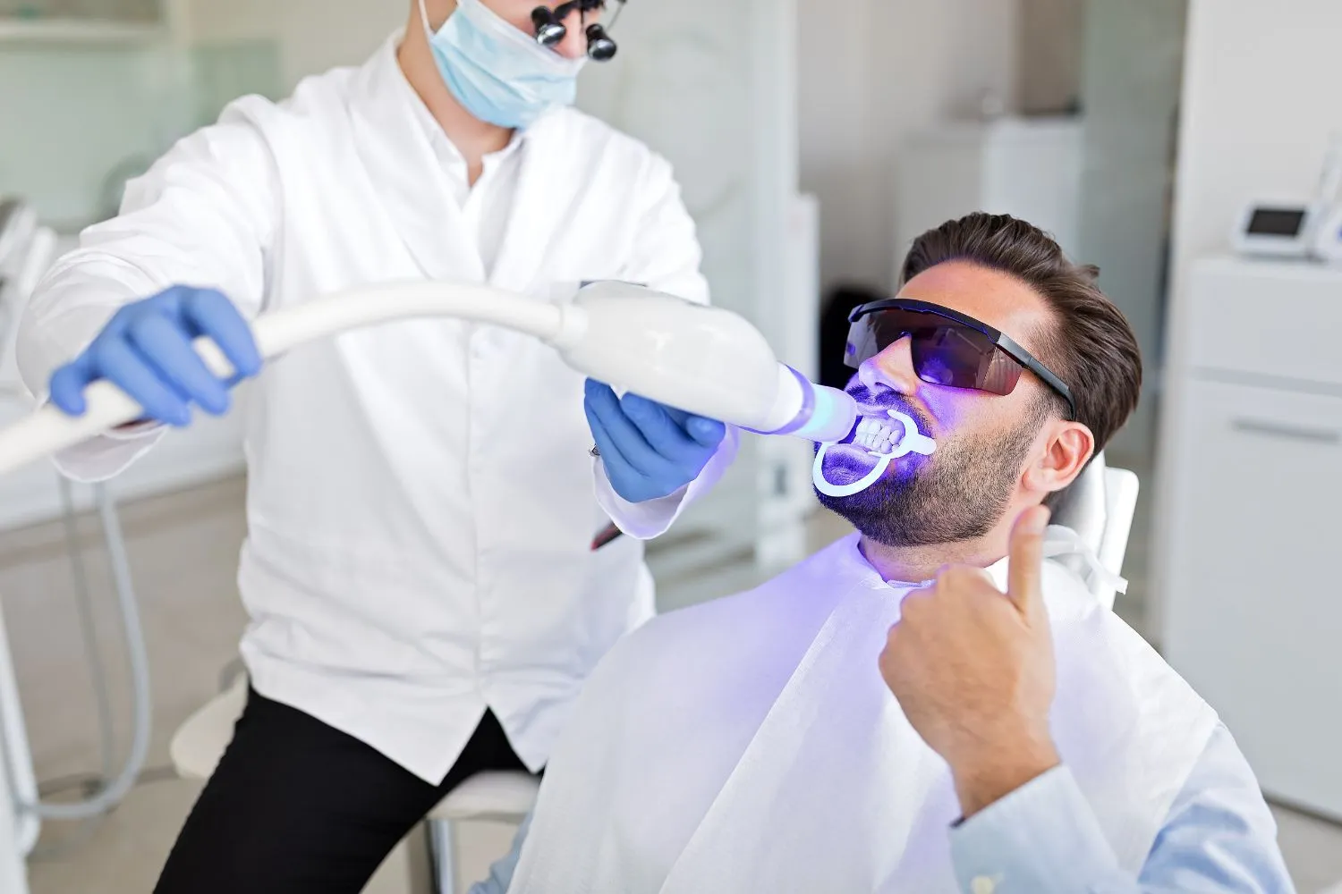 A male patient undergoing professional teeth whitening with a UV light while wearing protective eyewear, giving a thumbs-up in the dental chair.