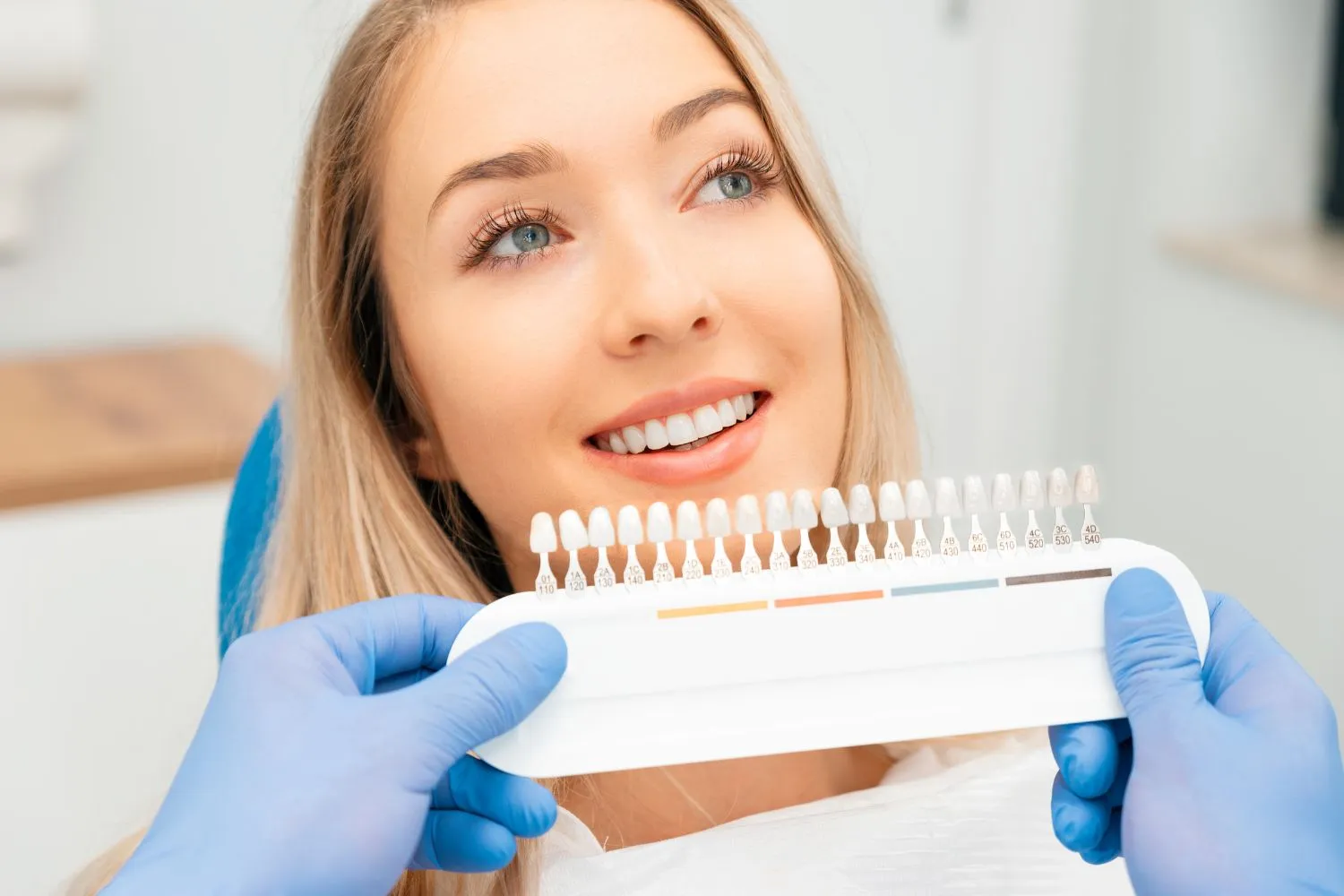 A happy blonde woman in the dental chair looking at a shade guide held by a dentist for tooth color matching.