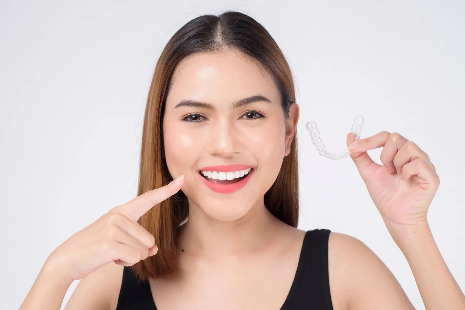 A woman smiling confidently while pointing to her teeth and holding a clear aligner, highlighting orthodontic treatment success.