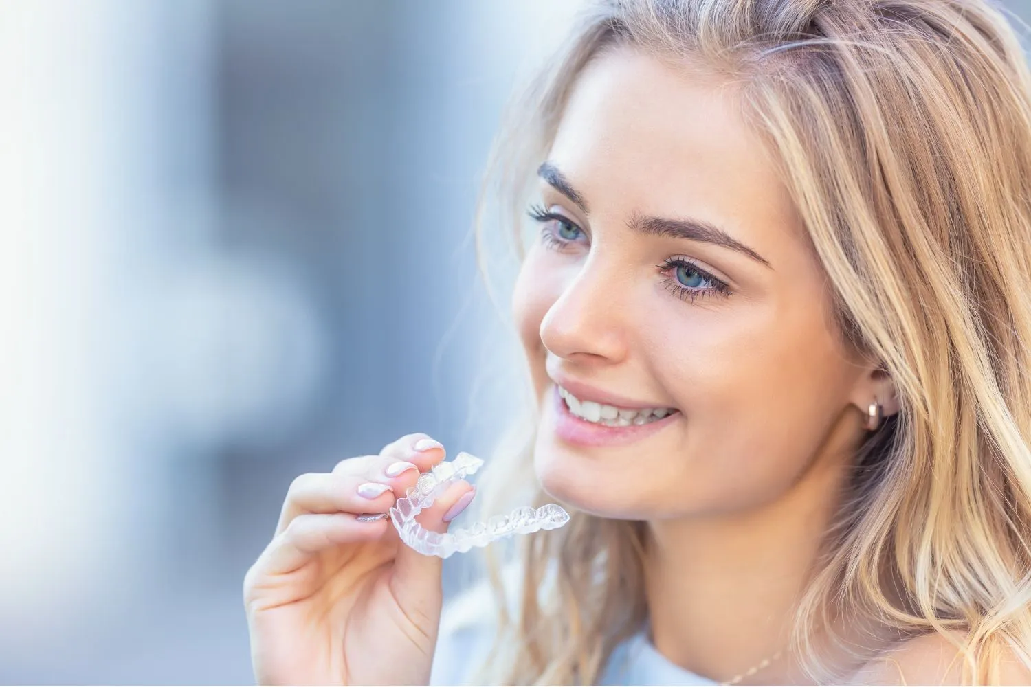 A blonde woman smiling while holding a clear aligner near her mouth, demonstrating modern orthodontic treatment.