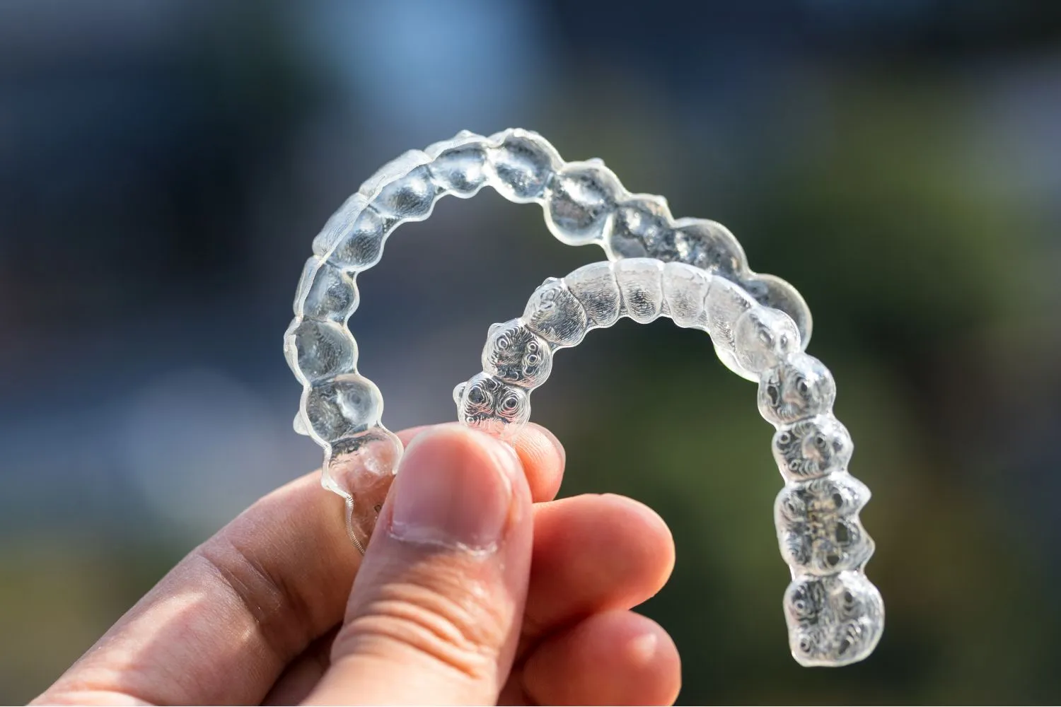 Close-up of a hand holding a transparent clear aligner with a blurred outdoor background, showing the orthodontic appliance.