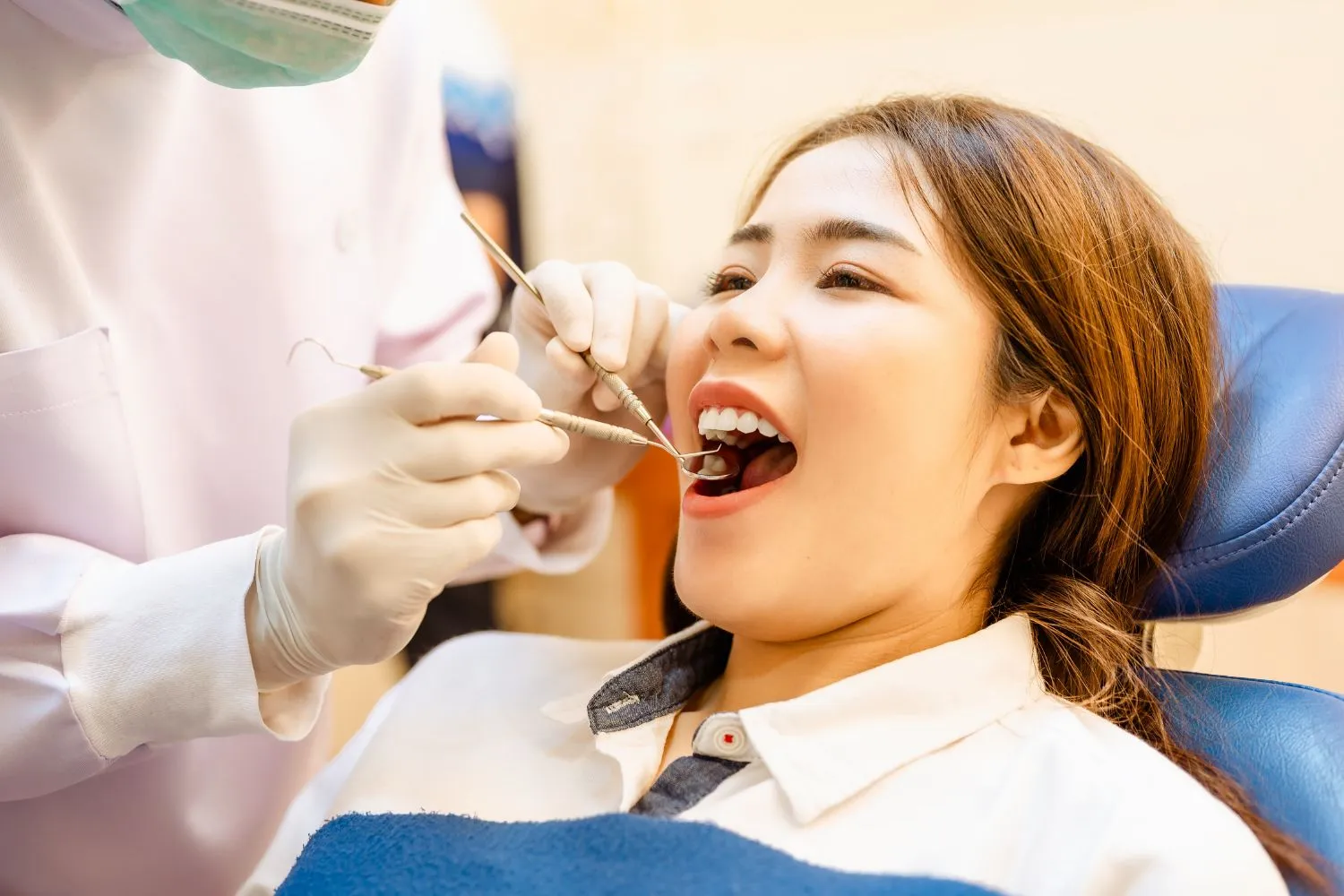 A woman sitting in a dental chair with her mouth open as a dentist examines her teeth using a dental mirror and explorer.