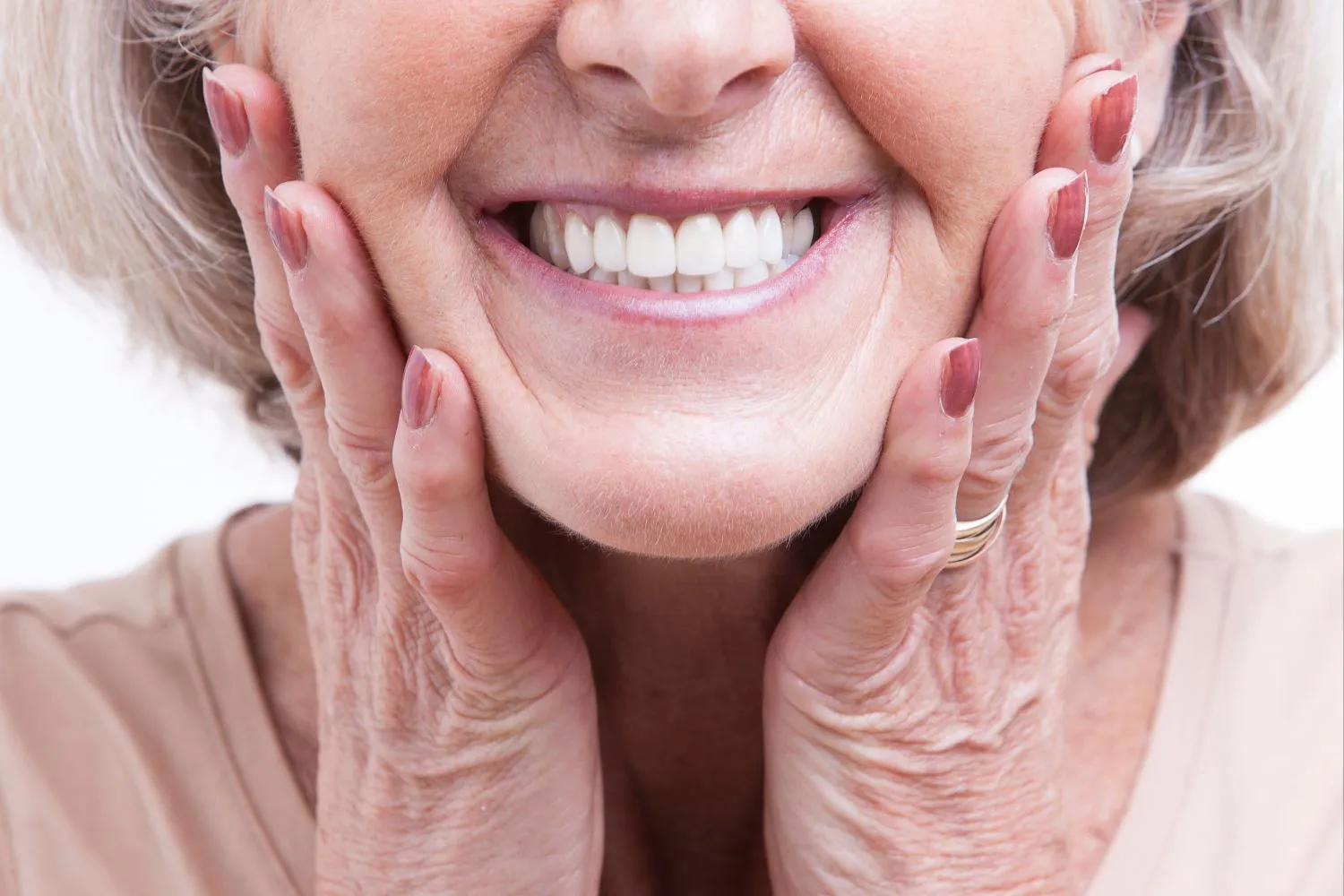 Close-up of a happy senior woman showing off her white dentures, smiling confidently with hands on her cheeks.