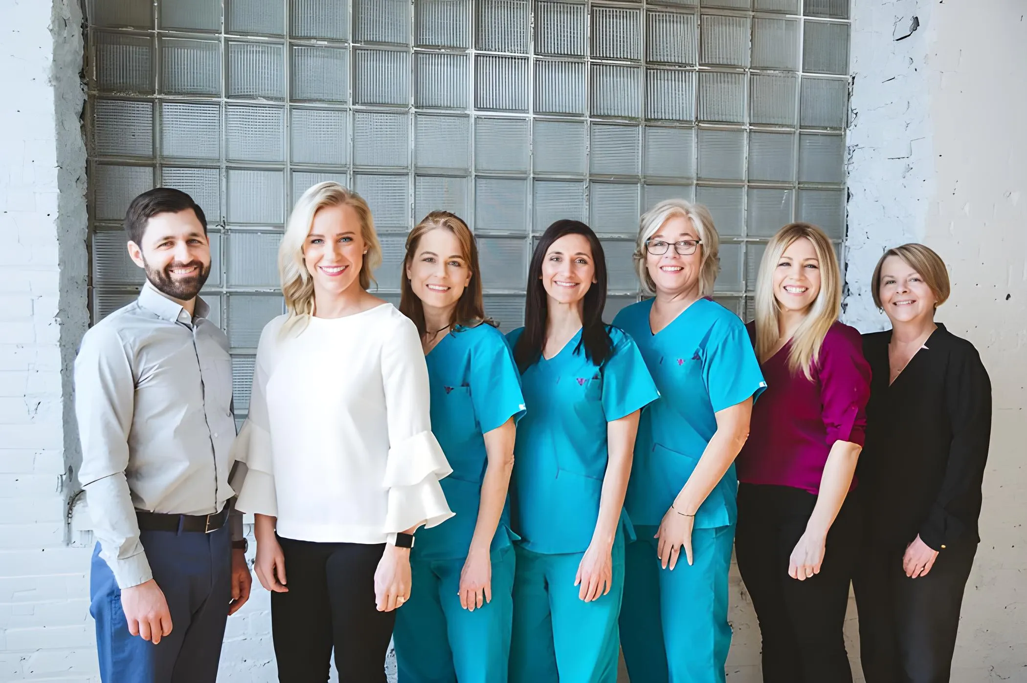 The Bates Dental Care team smiling together in front of a glass-block wall, featuring staff members in scrubs and business casual attire.
