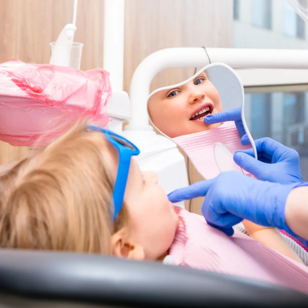 A young girl wearing blue glasses pointing at her reflection in a dental mirror during her appointment.