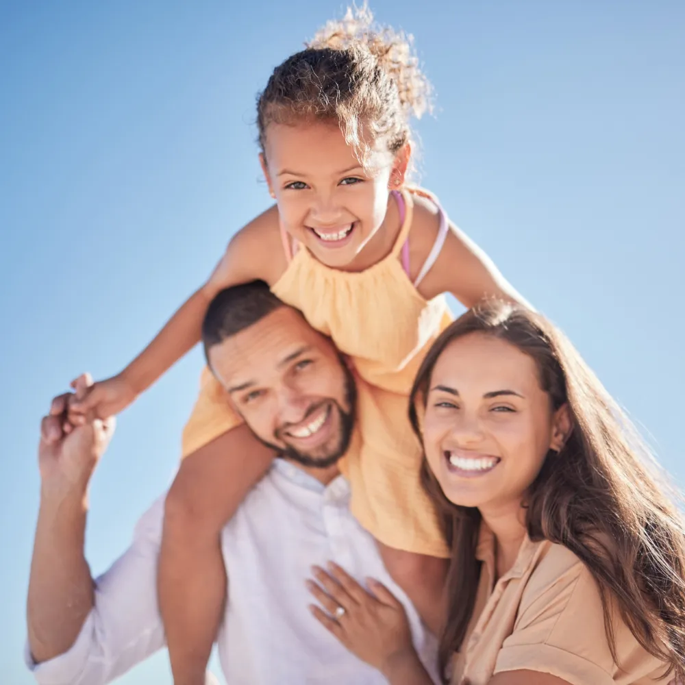 A joyful family of three posing outdoors on a sunny day, with a little girl riding on her father's shoulders.