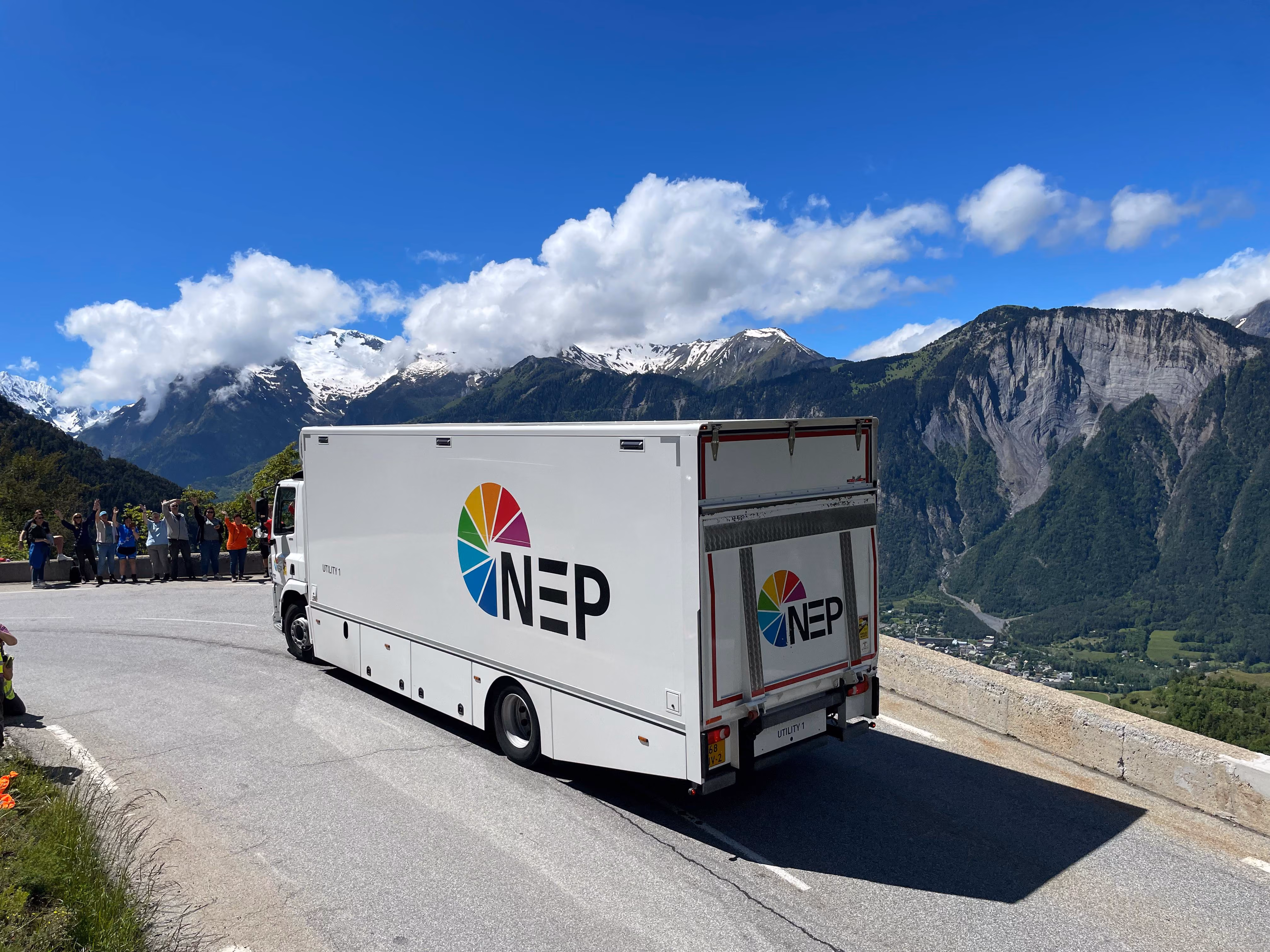 White NEP truck driving on curvy mountain road with snowy Alps and spectators in background.