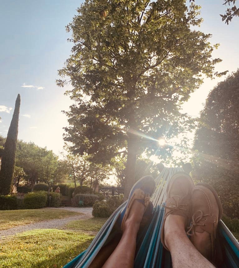 Two pairs of feet in casual shoes rest in a striped hammock, overlooking a serene Italian garden with lush greenery, a gravel path, and sunlight filtering through tall trees.