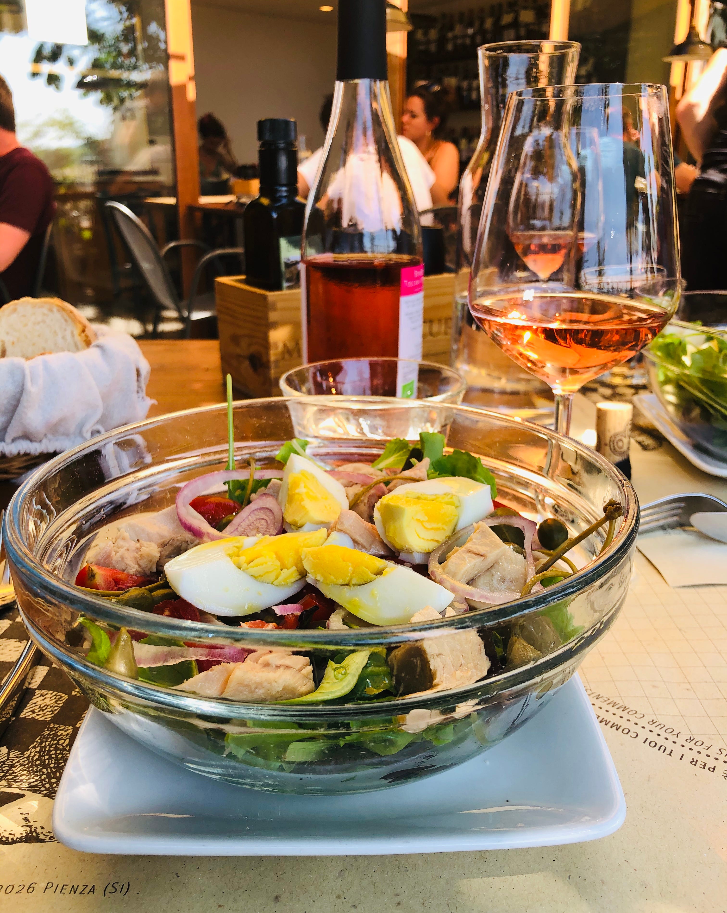 A glass bowl of fresh salad with tuna, boiled eggs, red onions, and greens sits on a rustic table, accompanied by a glass of rosé wine, a bottle of wine, and a basket of bread in a cozy Italian restaurant setting.