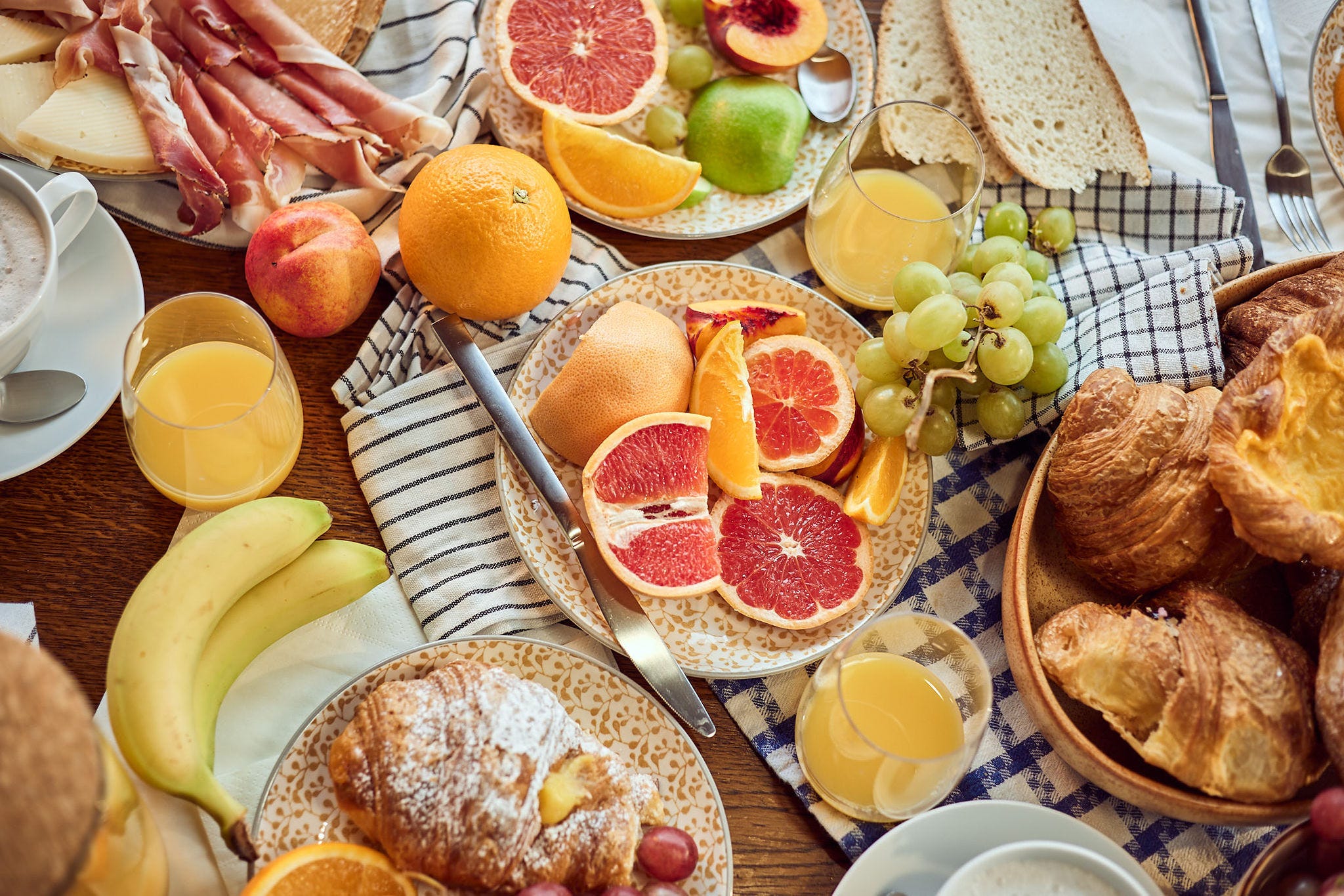 A rustic table is laden with an Italian breakfast spread, including fresh oranges, grapefruits, grapes, croissants, charcuterie, and glasses of orange juice, all arranged on a checkered cloth with a cup of coffee.