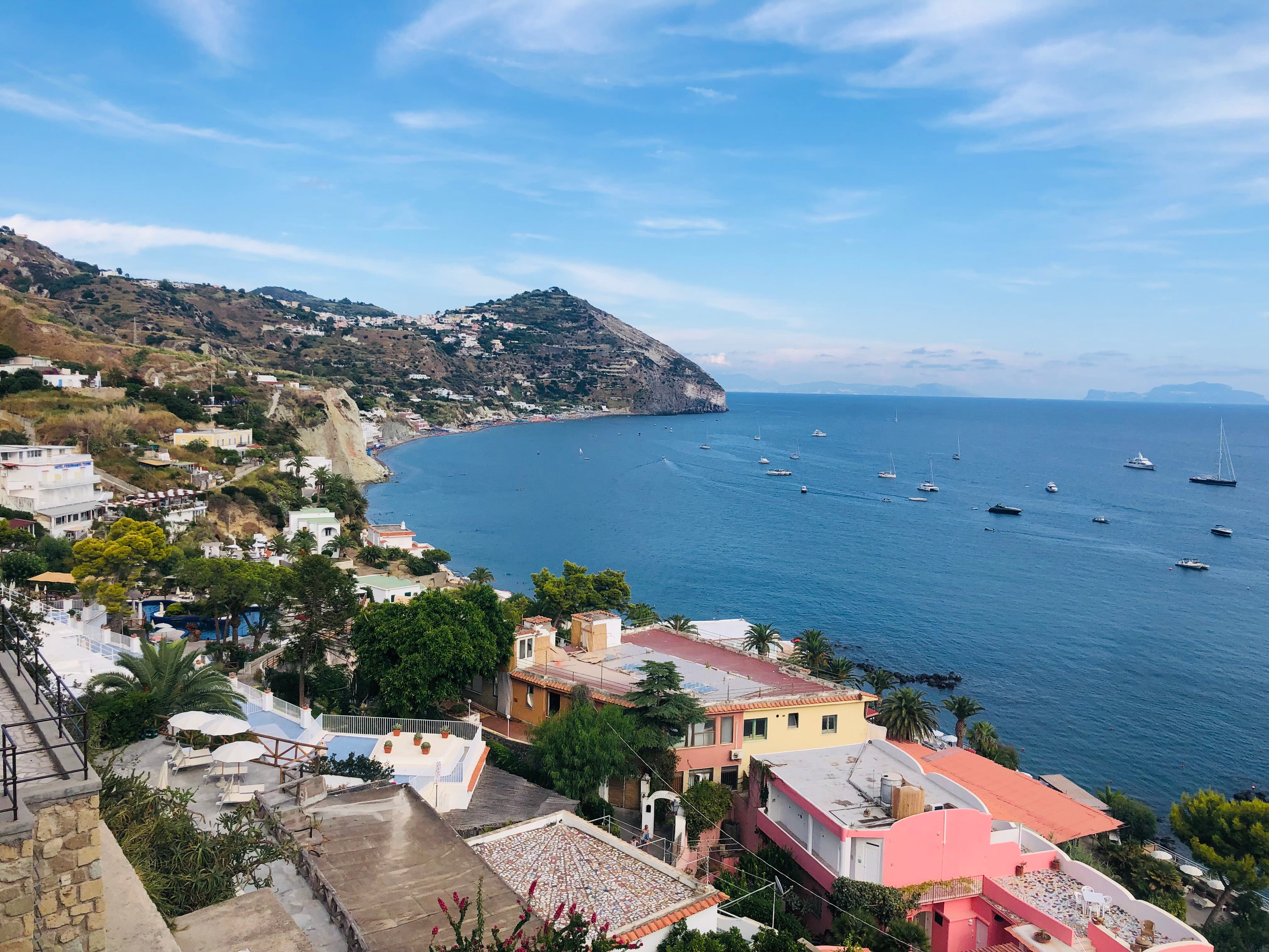 A scenic view of an Italian coastal village with colourful buildings, palm trees, and a clear blue sea dotted with boats, framed by rugged cliffs and a bright sky.