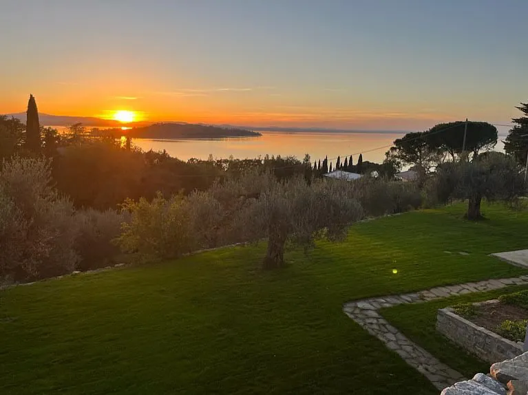 A vibrant sunset casts an orange glow over a peaceful Italian landscape, featuring olive trees, a well-kept lawn, and a distant view of a lake or sea with hills on the horizon.