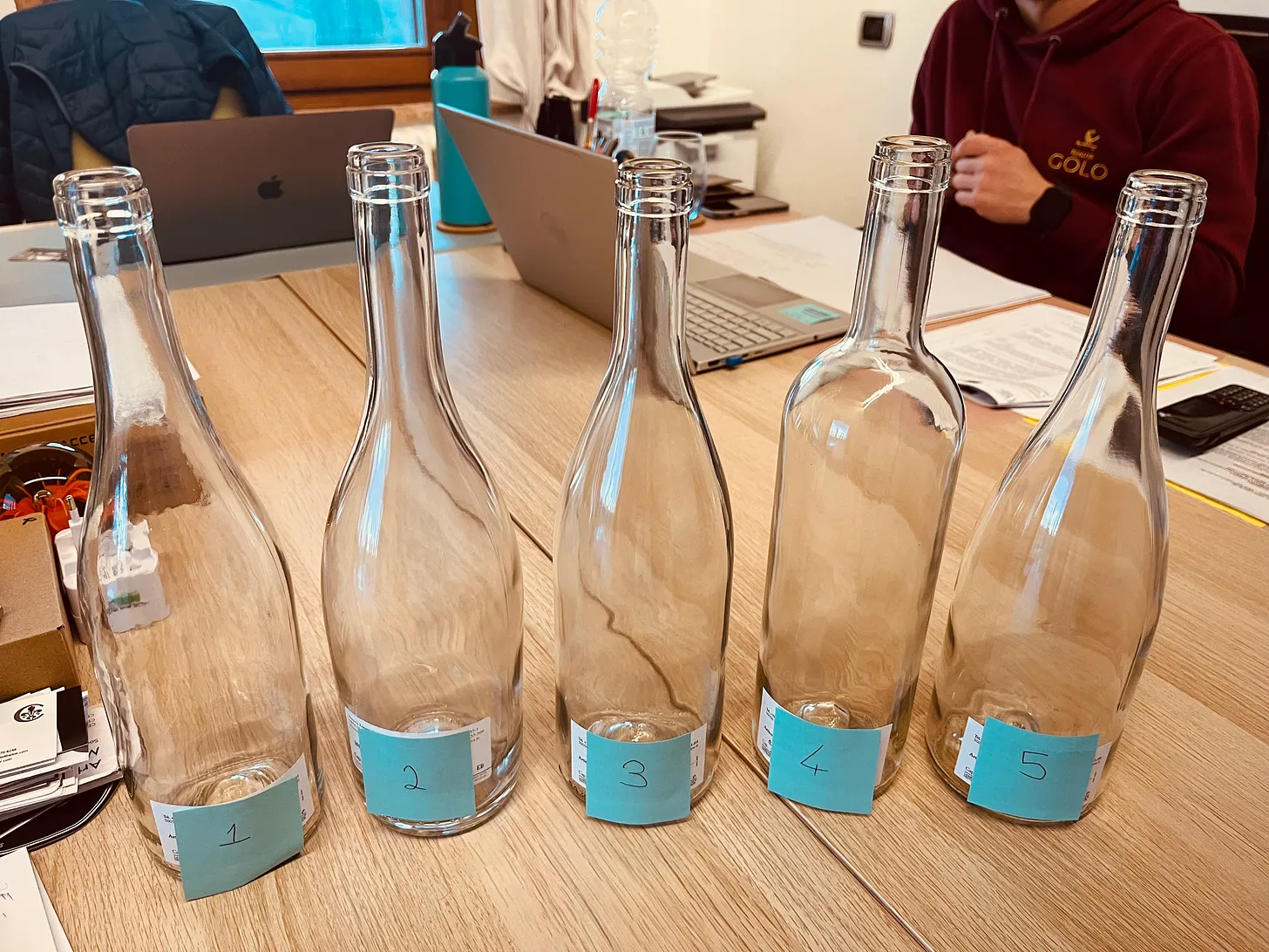 Five empty wine bottles labelled 1 to 5 on a wooden table during a bottle selection meeting at Tenuta Golo Winery in Montepulciano, with laptops and paperwork in the background.
