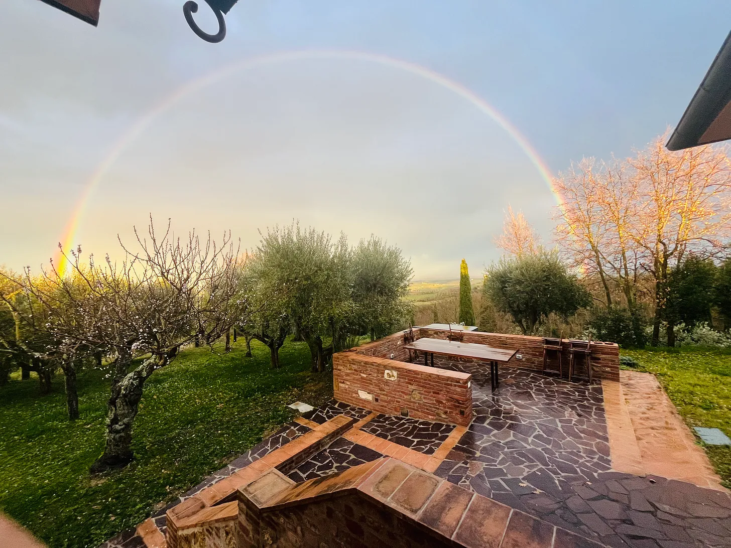 A rainbow arches over the outdoor patio at Tenuta Golo Winery in Montepulciano, with a rustic brick table and olive trees in the lush landscape at sunset.