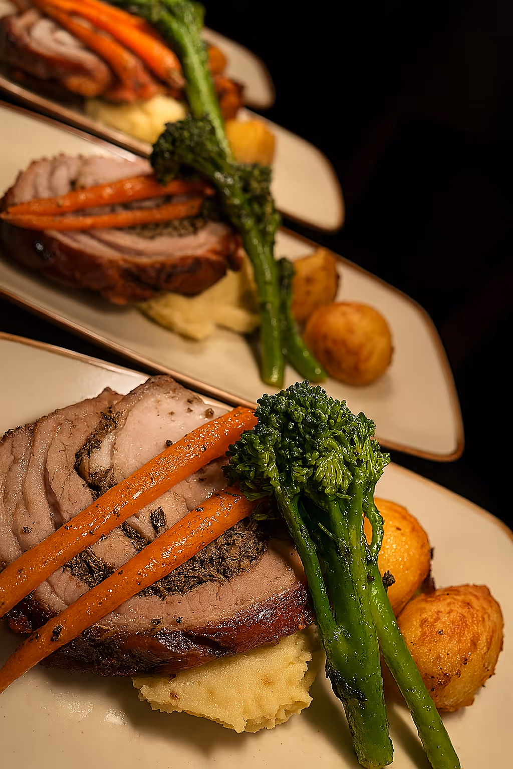 Plates of sliced roast beef served with roasted carrots, broccolini, two roasted potatoes, and mashed potatoes.