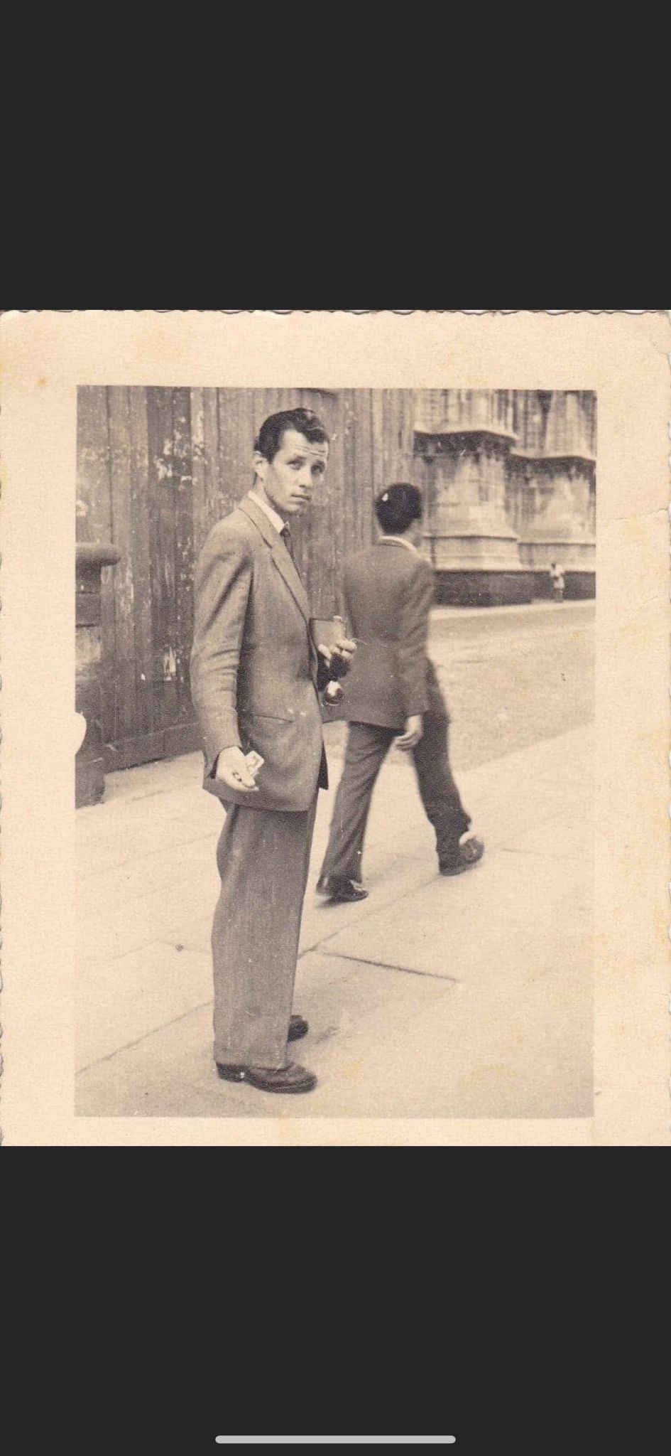 Black and white photo of a man in a suit holding a drink and a cigarette, standing on a sidewalk with another man walking away.
