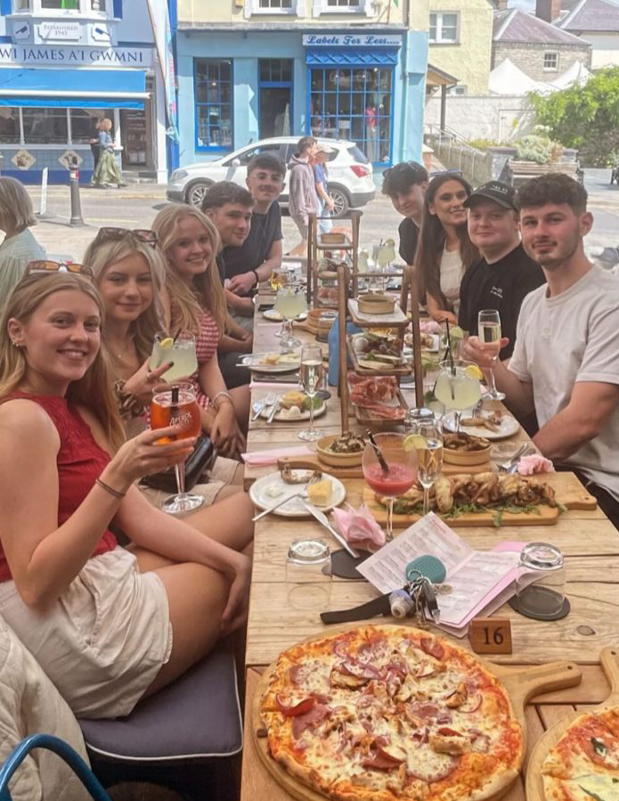 Group of nine young adults sitting at a long outdoor table enjoying food and drinks including pizza, cocktails, and appetizers.
