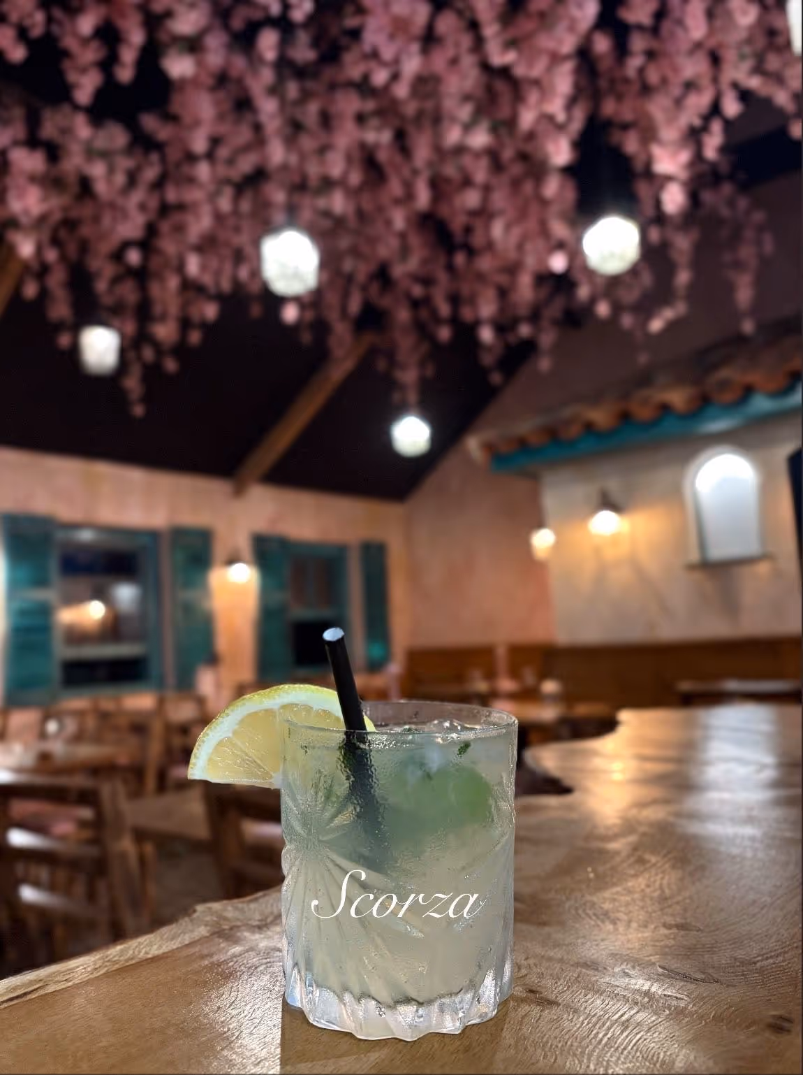 Iced cocktail garnished with a lemon slice on a wooden bar inside a cozy restaurant with hanging pink flowers and warm lighting.