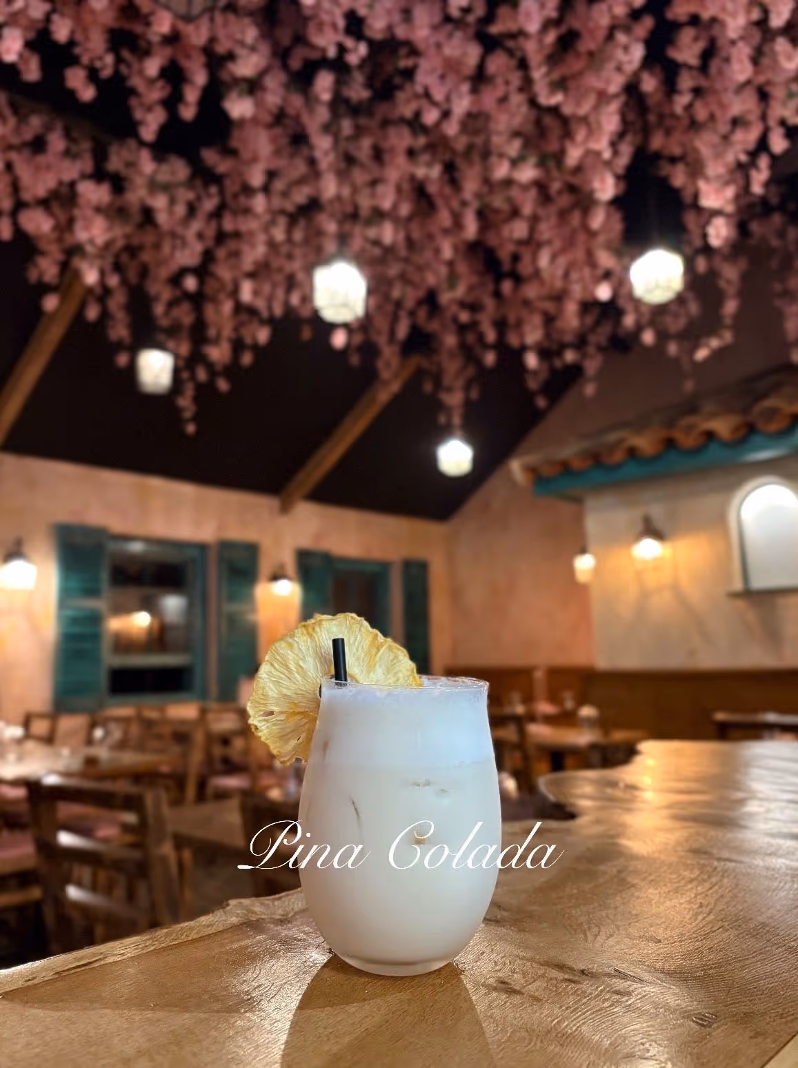 Pina colada cocktail garnished with a dried pineapple slice on a wooden table in a cozy restaurant with pink floral ceiling decorations and warm lighting.