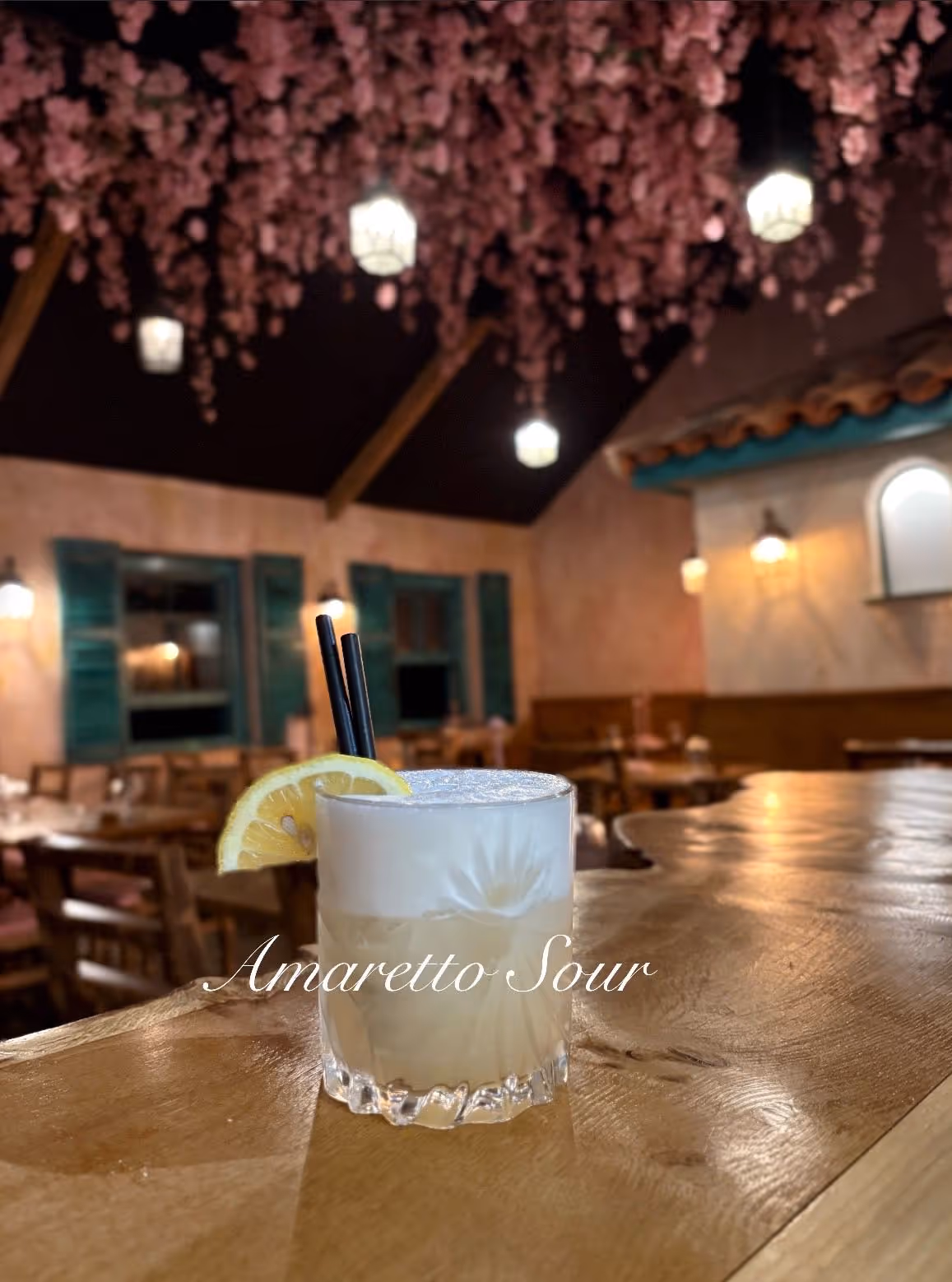 Amaretto Sour cocktail with lemon garnish and two black straws on a wooden bar with pink flower decorations and cozy restaurant interior.