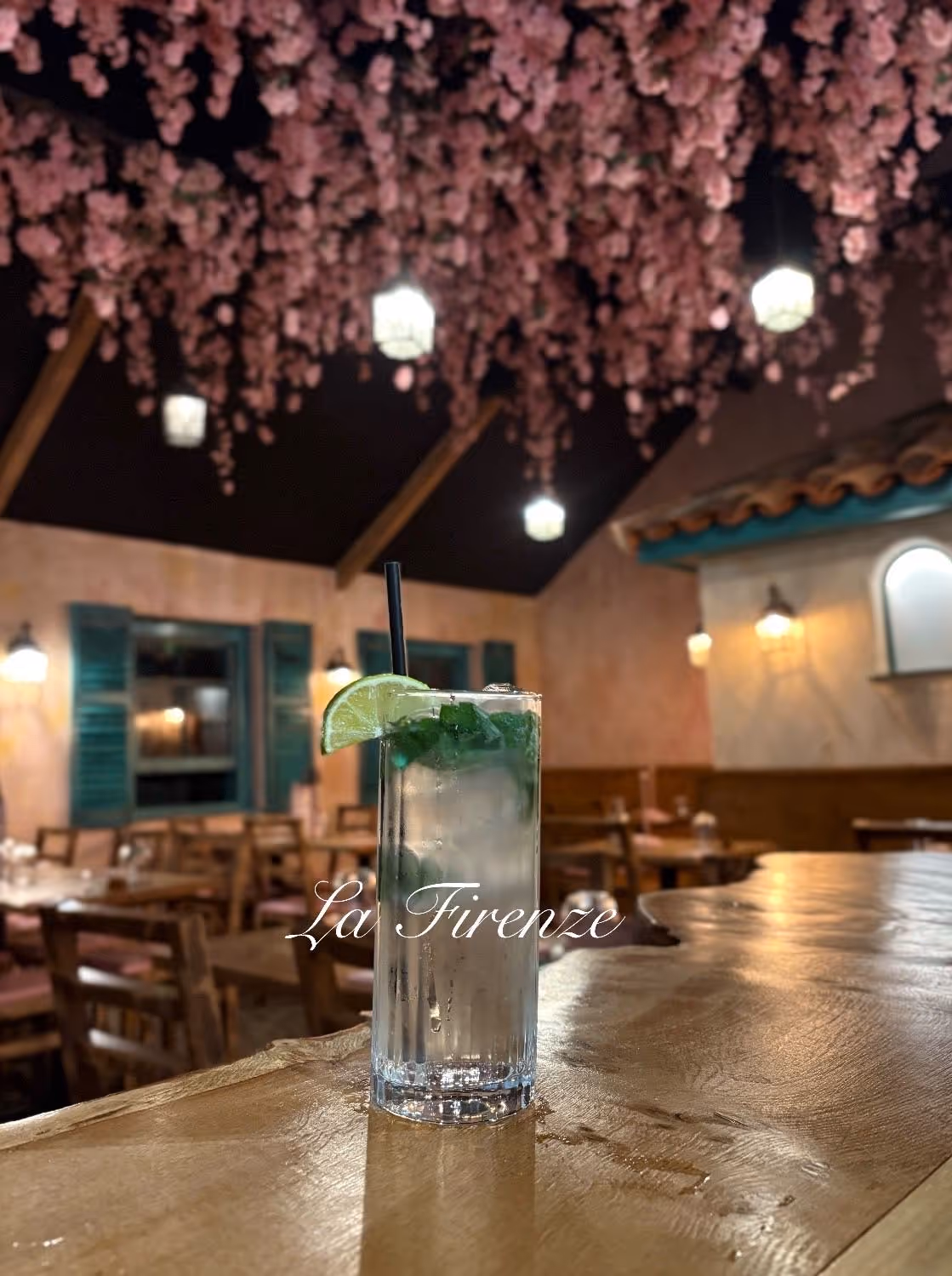 Tall glass of iced drink garnished with lime and mint on a wooden bar with cozy indoor seating and hanging pink flowers above.