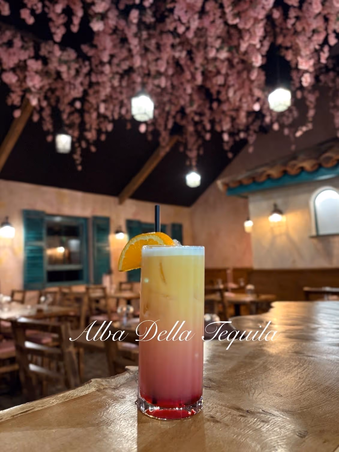 Layered tequila sunrise cocktail with an orange slice garnish on a wooden bar, under hanging pink flowers and warm lights in a cozy restaurant.