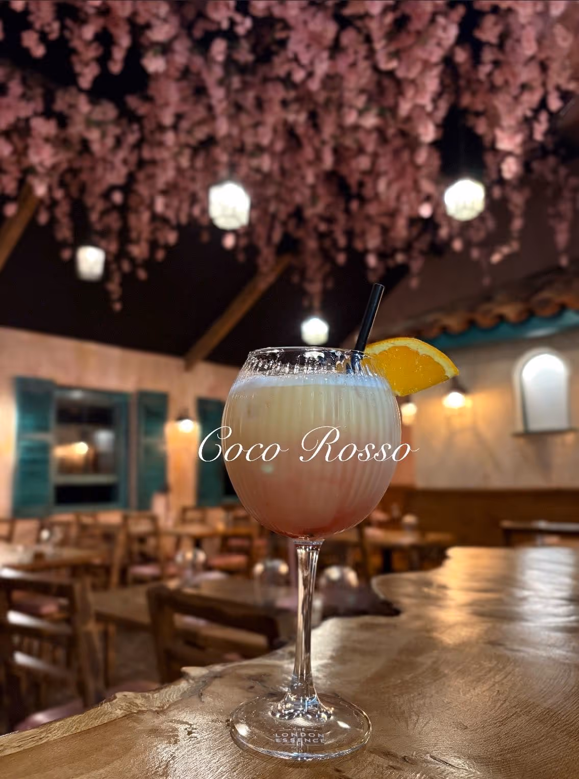 Cocktail glass with a creamy pink drink garnished with an orange slice, on a wooden bar counter in a cozy restaurant with pink hanging flowers and warm lighting.