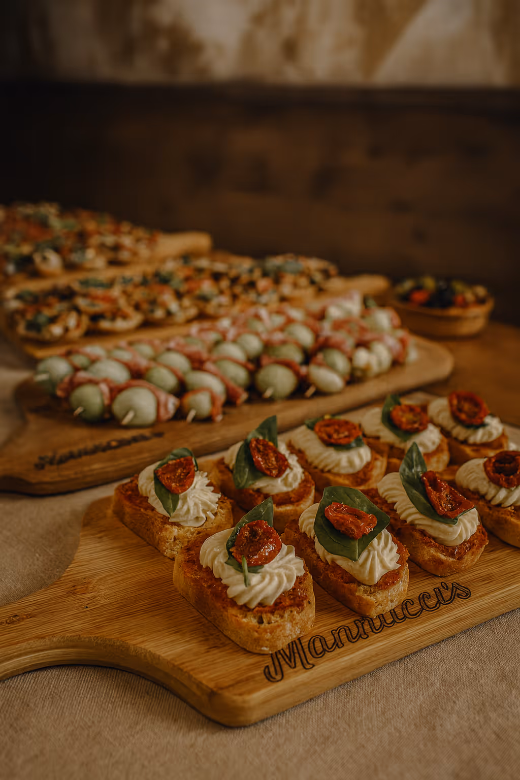 Wooden boards with assorted gourmet appetizers including crostini topped with cream, a basil leaf, and sun-dried tomato, and skewers of mozzarella and cured meat.