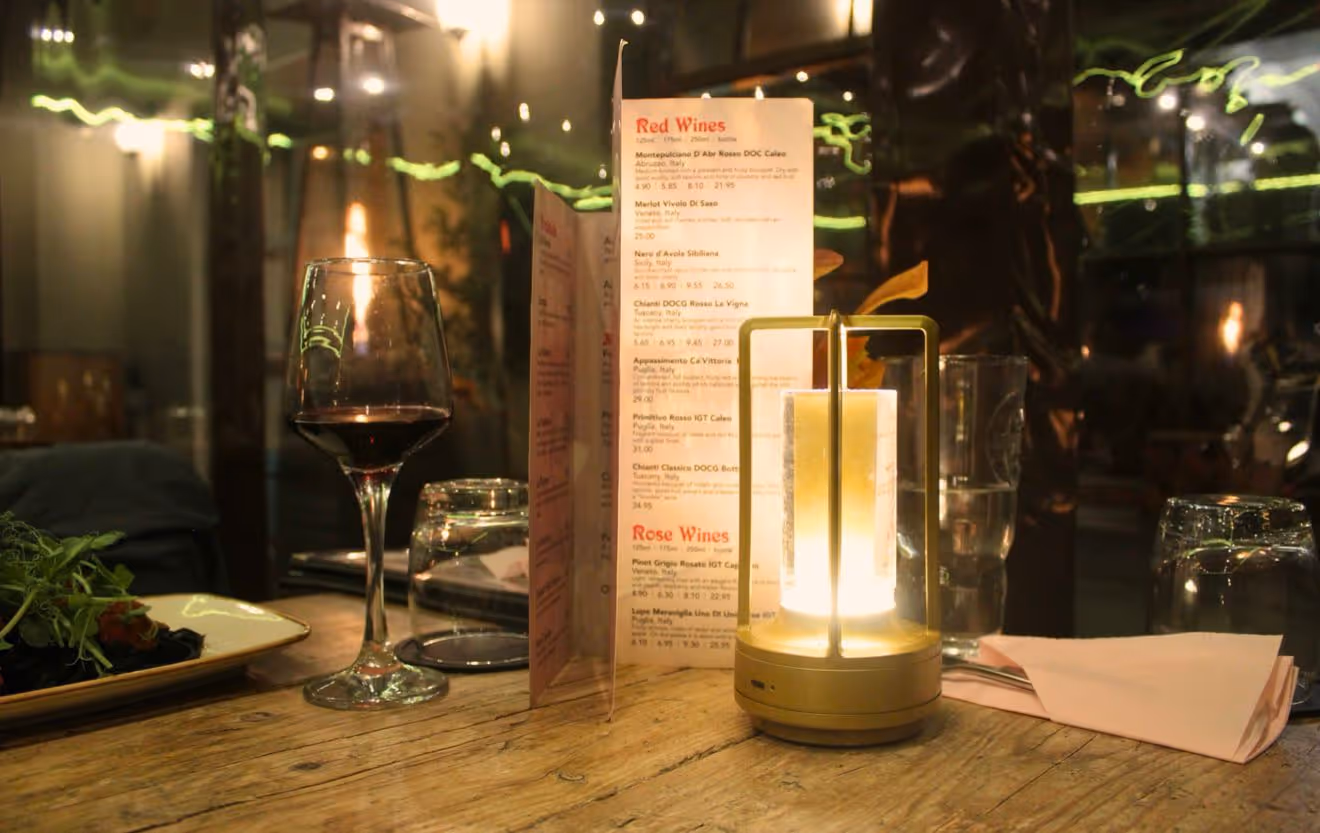 Wooden table at a dimly lit restaurant with a glass of red wine, a menu, a modern candle lantern, glasses, and a plate of salad.