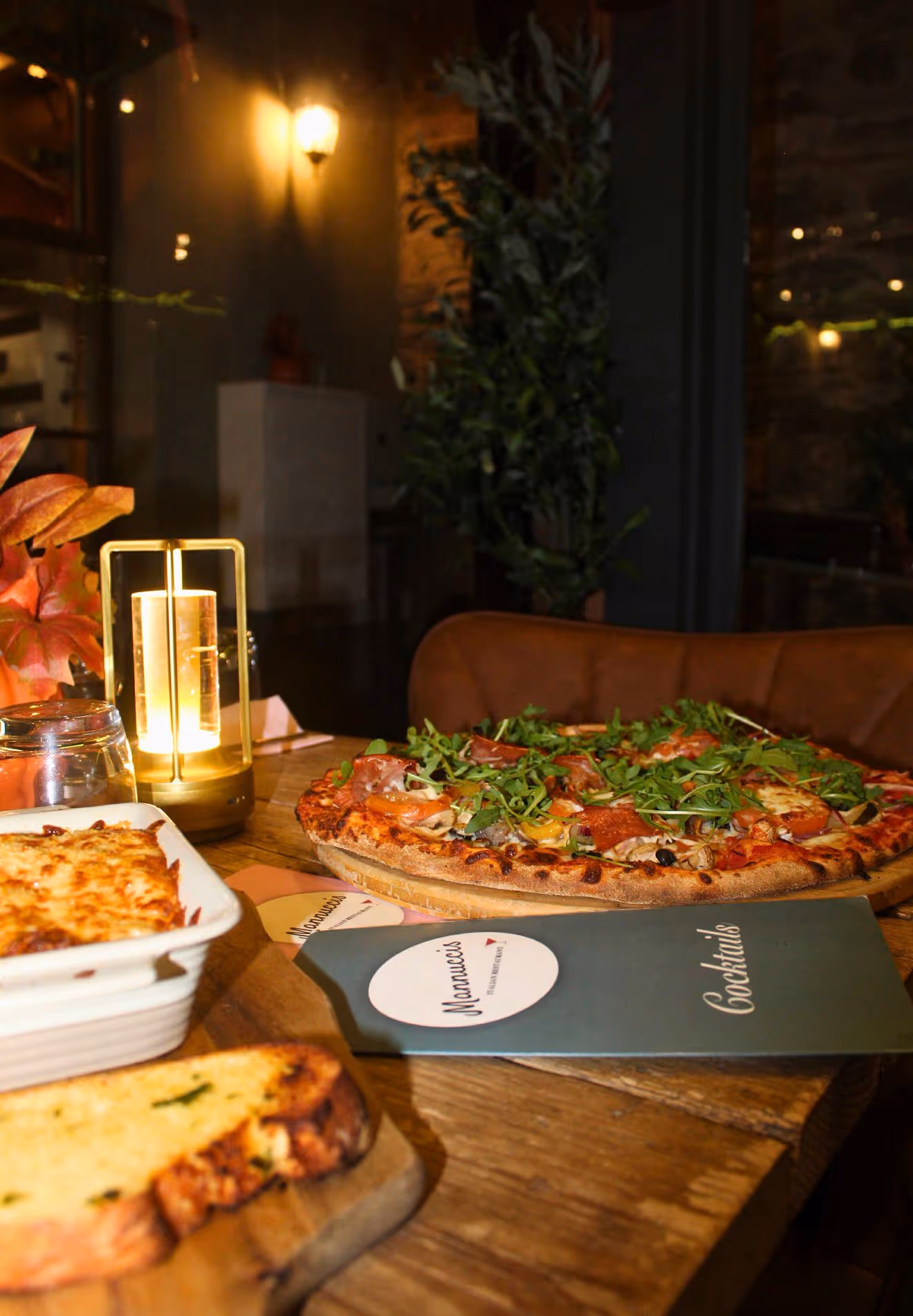 Table in a dimly lit restaurant featuring a pizza topped with arugula, a glass, a warm bread slice, a baked pasta dish, and a cocktails menu.